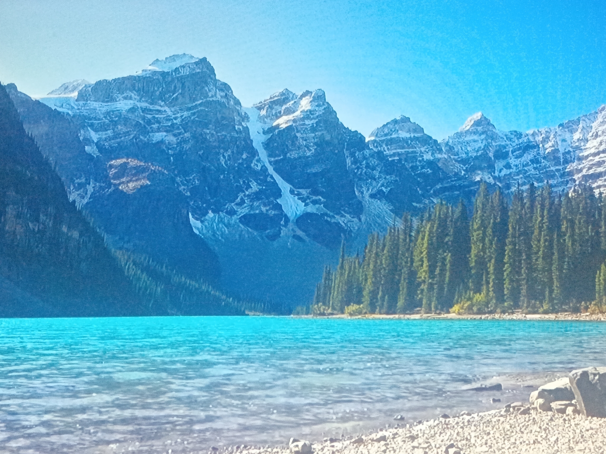 The image depicts a scenic view of a turquoise lake surrounded by snow-capped mountains and lush evergreen forests. The foreground features a shoreline with pebbles. The sky is a clear, bright blue. The landscape suggests a mountainous or alpine region. This image is not located in Juan Griego, Venezuela.