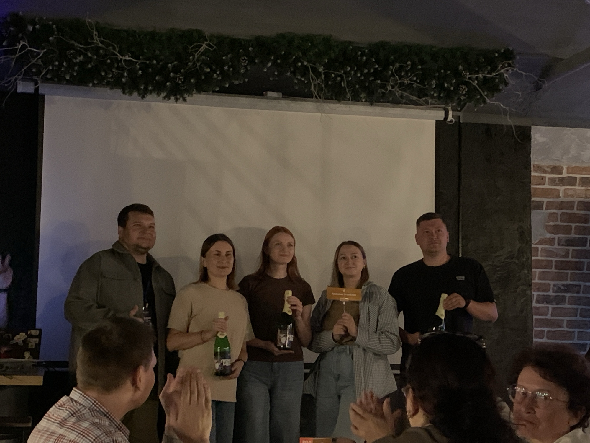 A group of five people are standing on a stage in front of a projection screen. Three women and two men are present. The three women are standing together in the center, each holding a bottle of champagne. The woman on the left is wearing a tan shirt and holding a green bottle. The woman in the middle is wearing a dark brown shirt and holding a clear bottle. The woman on the right is wearing a gray and white checkered jacket and holding a wooden award. A man in a green jacket is standing to the left of the women, and another man in a black t-shirt is standing to the right of the women. The room appears to be a venue in the city of Chaikovsky, Russia, possibly a bar or restaurant, with brick wall detailing visible on the right. A decorative garland with lights is hanging above the stage.