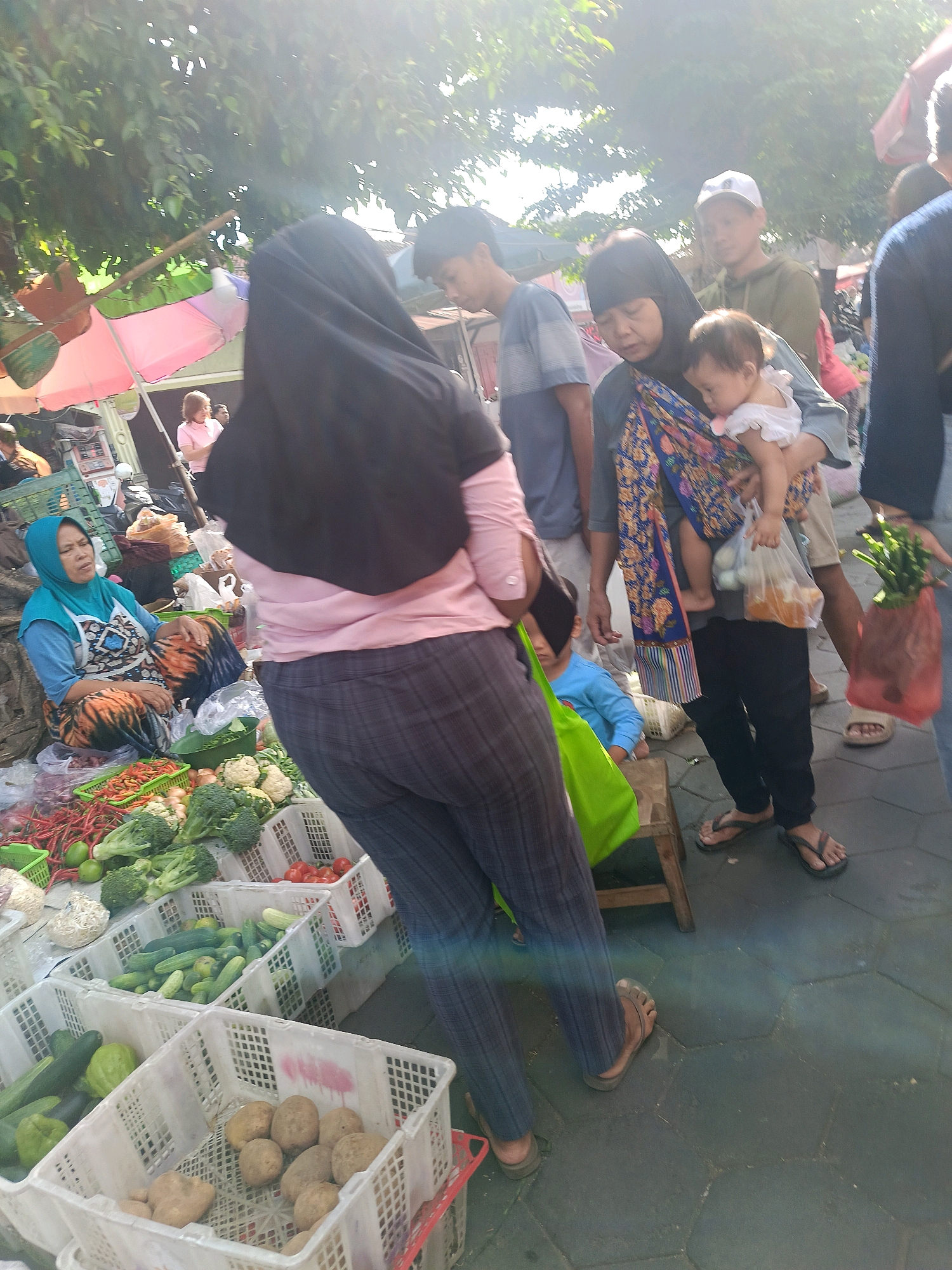 In Yogyakarta, Indonesia, a bustling street market scene unfolds under a bright sun. A woman wearing a black hijab and pink shirt is captured from behind, her patterned pants a prominent detail. To her left, a woman in a blue hijab and apron sits amidst a colorful array of vegetables, including red chilies, broccoli, and cauliflower, contained within white plastic crates.
Further into the scene, a woman with a dark hijab cradles a baby wrapped in a patterned cloth, her gaze directed downwards. She stands near a man wearing a white cap and olive-green hoodie, who appears to be looking in the same direction. Another man, wearing a blue striped t-shirt, is also visible in the background, as are several market stalls with umbrellas providing shade.
The ground is paved with hexagonal tiles, reflecting the sunlight. Bags of produce and other market goods are scattered throughout the scene, indicating a lively commercial environment. The overall impression is one of everyday life in a vibrant Indonesian market.