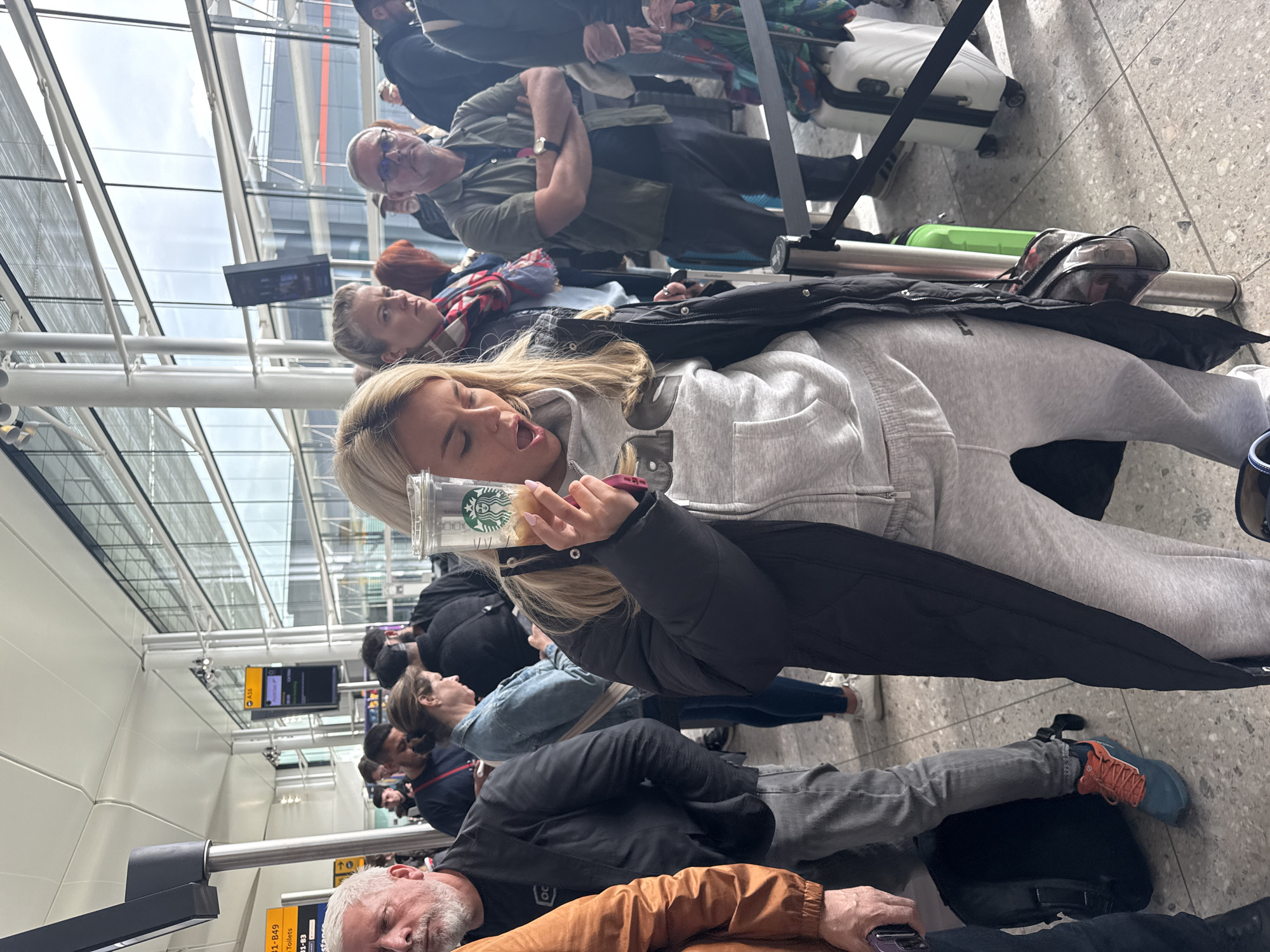 The image depicts a crowd of people waiting in a queue inside an airport terminal, presumably in London, United Kingdom. The terminal has a high ceiling with visible structural supports and large windows.
The people appear to be a diverse group, waiting with their luggage. They are standing behind guide rails. Some are holding coffee cups, and some are looking at their cell phones.