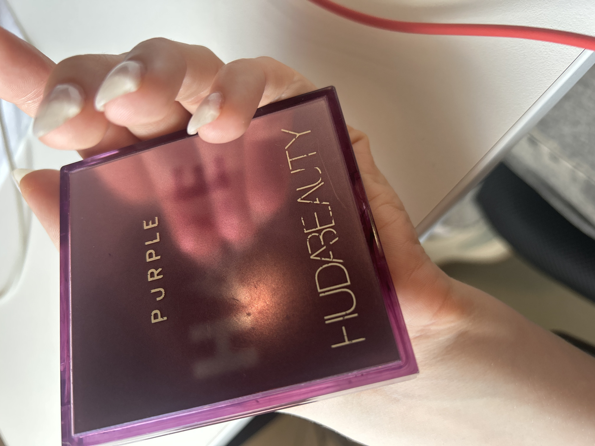 A person's hand with light skin and light-colored, almond-shaped fingernails holds a square, plum-colored makeup compact. The compact has gold lettering on its surface that reads "PURPLE" at the top and "HUDABEAUTY" at the bottom. The surface of the compact is reflective and shows a pinkish hue. The background is blurred and appears to be a white tabletop. The image's context within Perm, Russia is not determinable from the visual information provided.