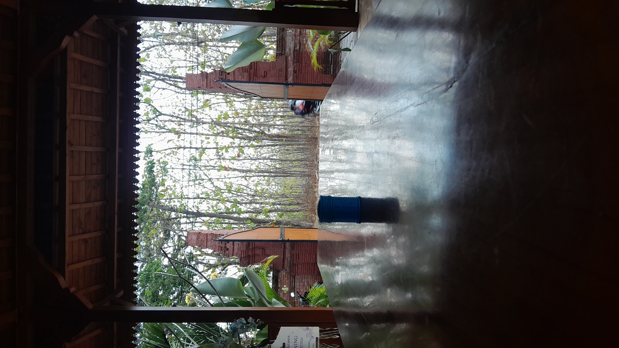 The image is a photograph taken from beneath a wooden pavilion, looking out towards a dense forest. In the foreground, a large, dark, polished wooden table reflects the surrounding environment. A small, cylindrical blue object, possibly a can or a container, sits on the table. In the middle ground, a red brick archway with wooden gates stands open, revealing the forest beyond. Through the archway, a person on a motorcycle is visible, driving away from the viewer. The forest consists of numerous trees with thin trunks and sparse foliage, suggesting a dry season. The sky is overcast. A sign with text is partially visible on the left side of the image. The scene appears to be an outdoor seating area, likely in a rural or natural setting. The location is stated as Giyanti, Indonesia.