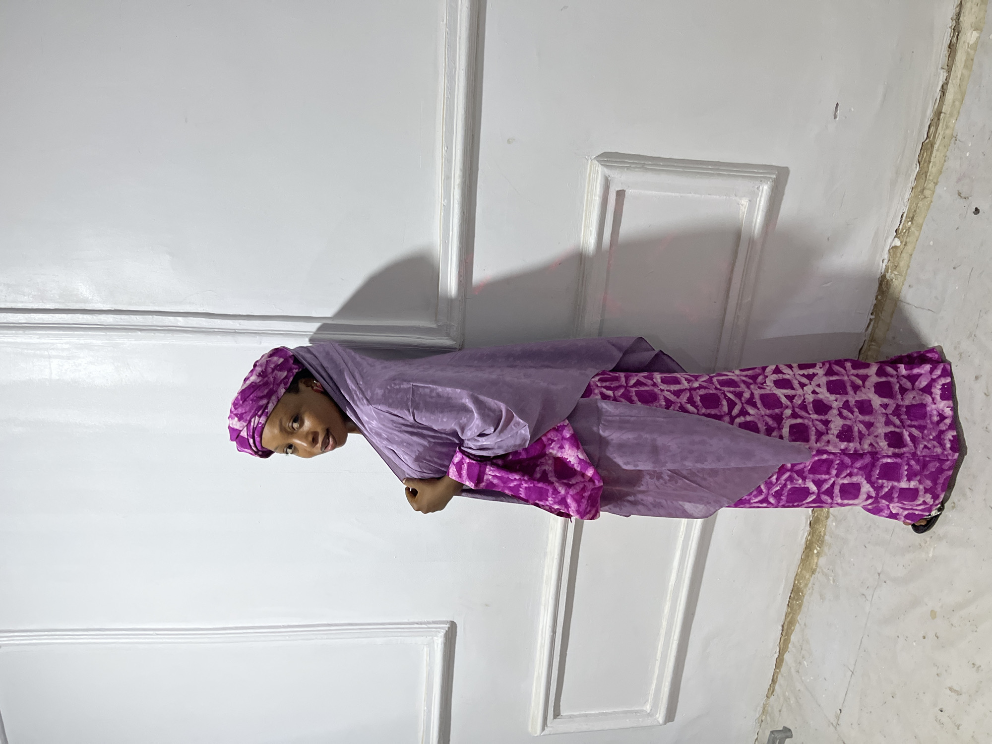 A woman in purple attire stands near a white wall with decorative moldings. Her clothing consists of a patterned skirt, a loose-fitting blouse, and a head covering. The location appears to be indoors, with a tiled floor visible in the lower right corner. The environment is minimally furnished. The location is not discernible as Gura Top, Nigeria, based on the image alone. The subject is facing left.