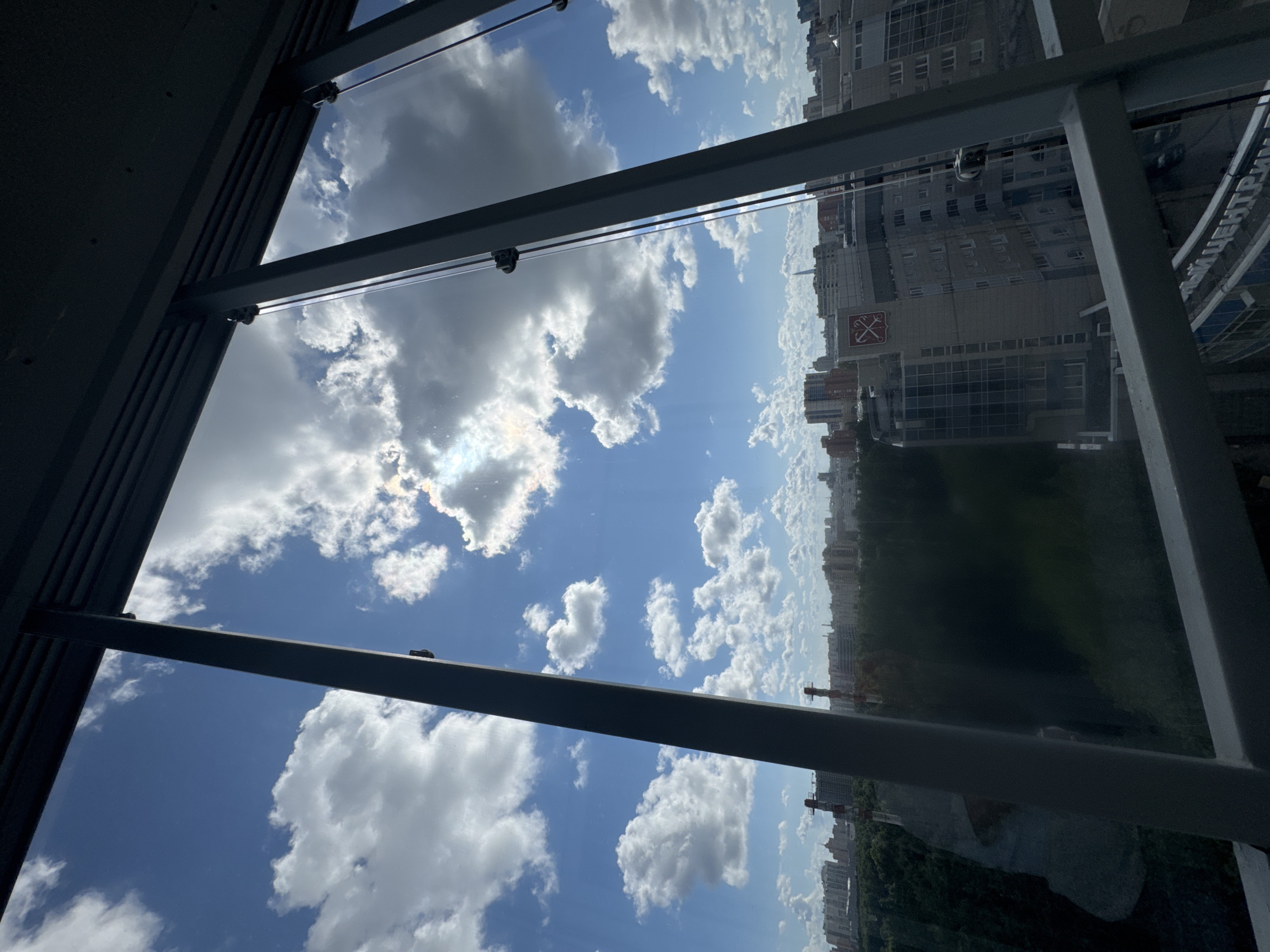 The image captures a daytime view of a cityscape, likely Saint Petersburg, Russia, seen through a modern building's glass facade and metal framing. The sky is a vibrant blue, populated with fluffy white cumulus clouds, with the sun partially obscured, creating a lens flare effect. Below the sky, a dense urban landscape unfolds. In the mid-ground, various buildings of different architectural styles and heights are visible, including some with glass exteriors and a building featuring a red emblem with crossed axes. Dense green trees form a thick line of foliage in the foreground, partially obscuring some of the lower buildings and a road below. The framing of the image suggests an elevated perspective from within a structure that has large, vertical glass panels and dark metal structural elements. There are no people visible in the image.