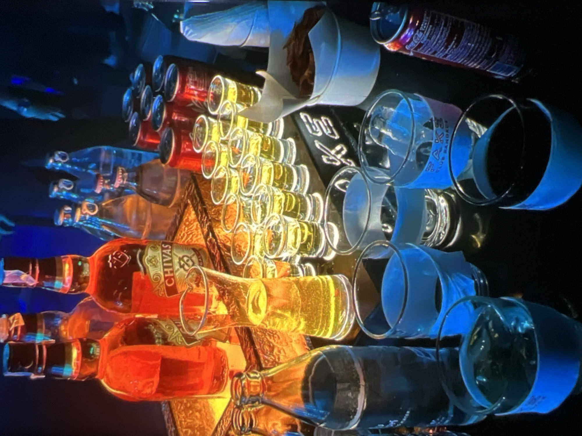The image depicts a bar or lounge setting, likely in Lagos, Nigeria, given the stylistic elements and potential cultural context. A prominent display features several bottles of alcoholic beverages, including what appears to be Chivas Regal Scotch whisky, alongside rows of shot glasses filled with a golden liquid.
In the foreground, there are several larger glasses, some containing ice and dark-colored liquid, possibly cola. These glasses are distinguished by blue bands around them, which appear to be illuminated. A tall, slender glass pitcher filled with a yellowish liquid is also visible.
To the right, a stack of red cans, resembling Coca-Cola, is present. Adjacent to the cans, a white cylindrical container holds a serving of what looks like fried snacks, possibly plantain chips or french fries.
The lighting in the scene is dim and atmospheric, with warm yellow and orange tones highlighting the bottles and shot glasses, and cool blue tones illuminating the foreground glasses and the "SAKE" lettering on the bar. The overall ambiance suggests a night-time establishment focused on drinks and socializing. There are no visible people in the frame, but the setup implies a service environment.