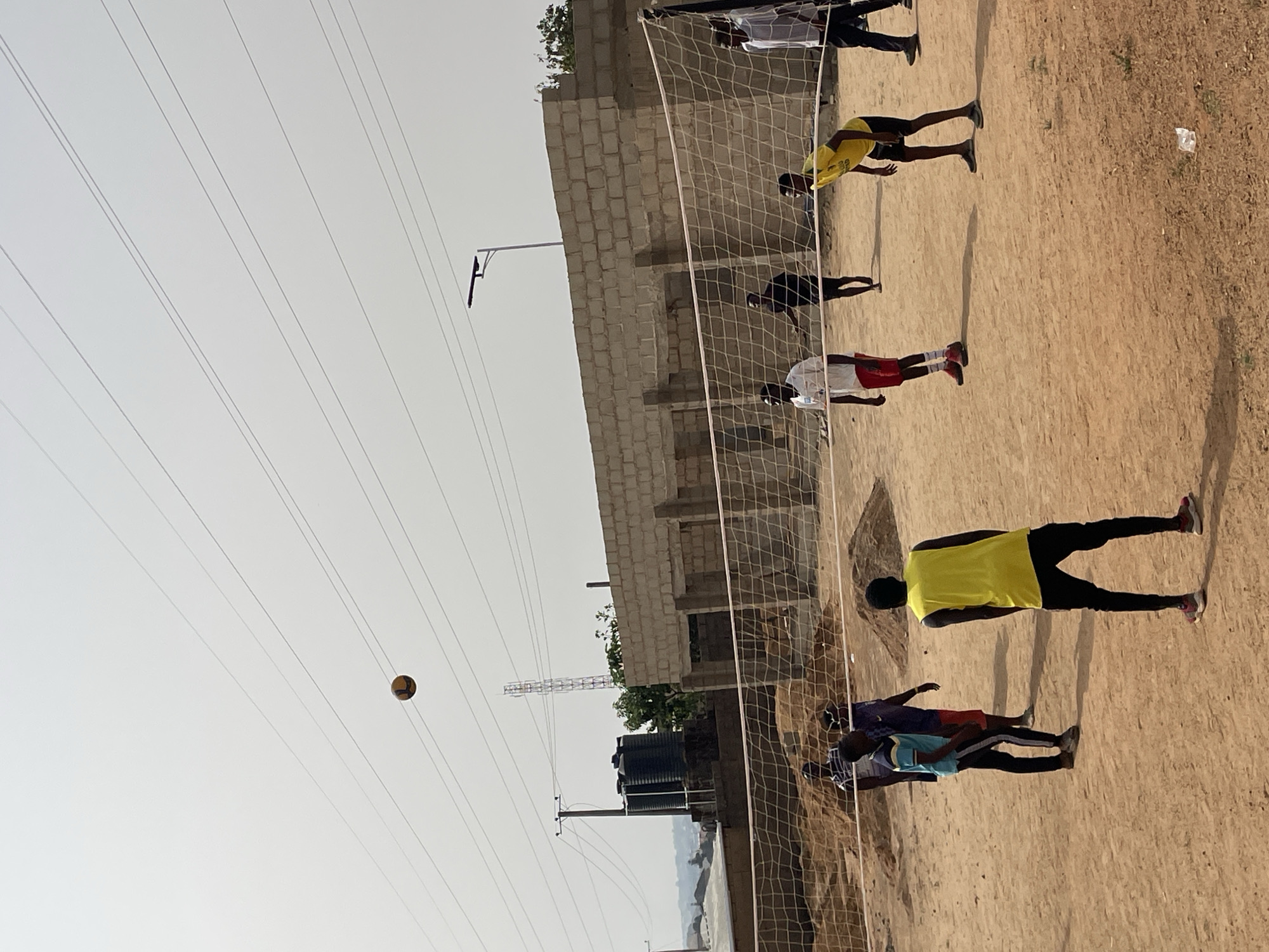 The image shows a group of people playing volleyball on a sandy court in Jos, Nigeria. A volleyball is suspended in the air against a pale sky with power lines. The volleyball net stretches across the court, with people in various athletic clothing positioned around it. A partially constructed brick building stands near the court, with water tanks and a communications tower visible in the background. The sandy surface of the court and the simple, unadorned environment suggest a casual, informal setting.