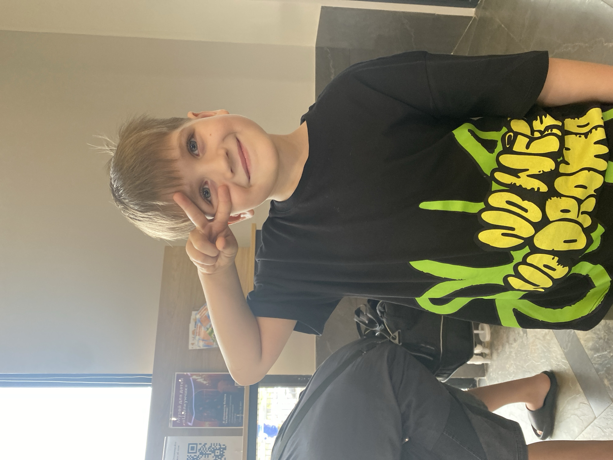 In Votkinsk, Russia, a young boy with blond hair and blue eyes, wearing a black t-shirt with a green and yellow graphic, is posing for a photo. He is holding up his right hand, forming a peace sign over his eye and smiling at the camera. He is standing indoors, possibly in a public building or hallway, with a window and a display of posters visible in the background. A person wearing a black shirt and gray shorts with dark sandals is partially visible to the left. Bags are also visible in the background.