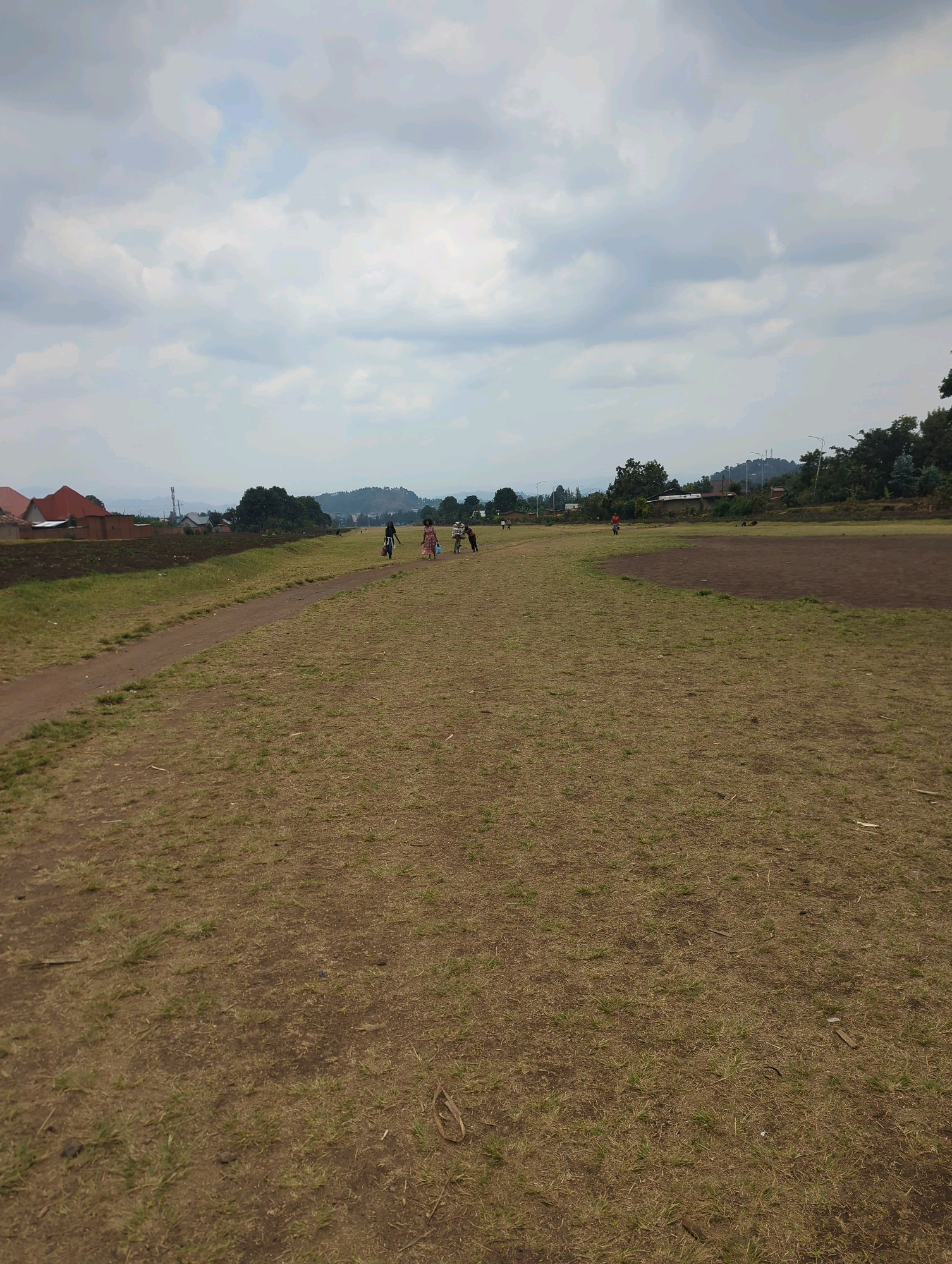 The image depicts an outdoor scene in the Musanze District of Rwanda, likely a field or open area on the outskirts of a settlement. The ground is covered in dry, light brown grass with patches of bare earth. A dirt path runs diagonally across the left side of the frame. In the background, there are several buildings with reddish-brown roofs, suggesting residential structures. Beyond the buildings, a line of trees forms a backdrop, and rolling hills or mountains are visible in the distance under a cloudy sky.
Several people are present in the scene, appearing to be children or young adults. On the right side of the dirt path, a group of four individuals are walking. One appears to be carrying a bag. Further into the field, to the left, another individual is visible, walking away from the camera. Another individual is seen in the far distance, further to the left. The overall atmosphere suggests a casual, everyday moment in a rural or semi-rural environment. The sky is overcast with a mix of light and dark grey clouds.