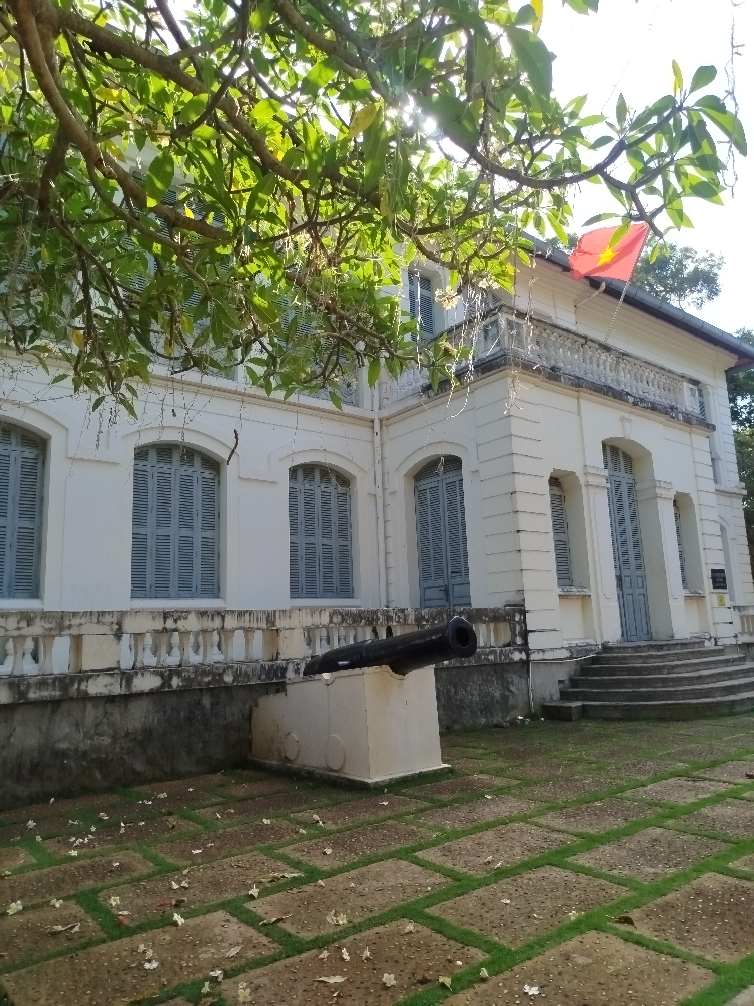 The image shows a white, two-story building in Ho Chi Minh City, Vietnam. The building has arched windows with blue shutters and a red flag on the roof. A cannon is positioned in front of the building, on a stone base. The ground is paved with stone squares separated by grass. Tree branches obscure the upper portion of the building.