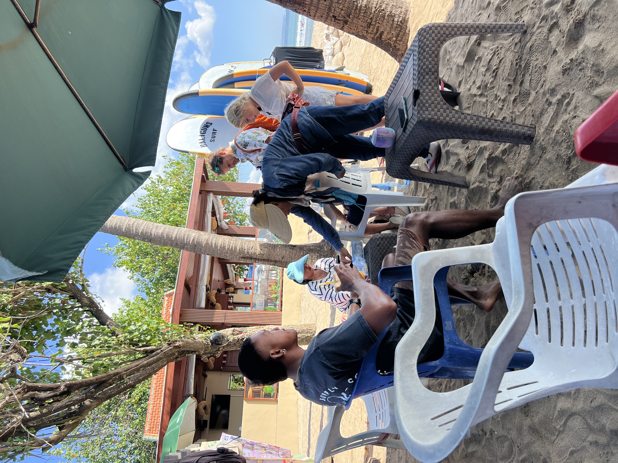 On a sandy beach in Kuta, Indonesia, under a large green umbrella, several people are gathered. In the foreground, a man with dark skin and short hair sits in a blue plastic chair, wearing a black t-shirt and patterned shorts. He holds a beverage. To his right, another person, partially obscured by a white plastic chair, is seated. In the middle ground, a group of individuals are interacting around a table. One woman wearing a hat and denim jacket leans forward, looking at something on the table. Another woman in a white shirt stands behind her, and a man in a floral shirt stands to the left. Behind them, two surfboards are propped upright. The background shows a beachside structure with a red-tiled roof, palm trees, and the ocean with small waves. The sky is blue with scattered clouds.