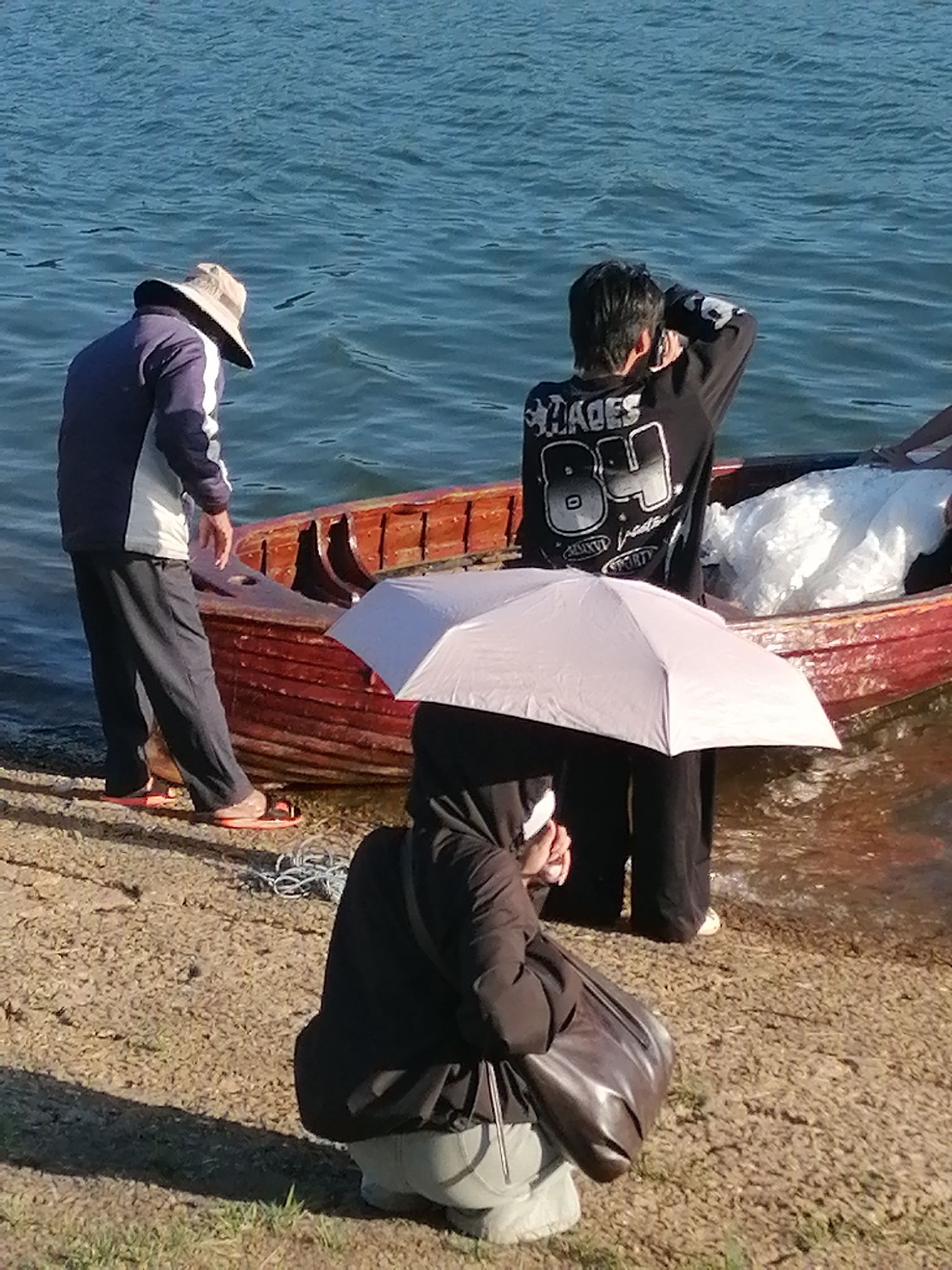 This image depicts a waterside scene in Da Lat, Vietnam, likely near Xuan Huong Lake, given the presence of a traditional wooden boat. The sunlight suggests it is daytime, possibly late afternoon, casting shadows. In the foreground, a person is squatting, holding a light pink umbrella, and wearing dark clothing and a light-colored hat. To the left, a person in a blue and white jacket and dark pants, wearing a wide-brimmed hat, stands near the water's edge. Behind them, a red wooden boat is partially submerged, with white material visible inside. Another individual, seen from the back, wears a black long-sleeved shirt with a graphic design and stands near the boat, looking out towards the water or engaged with something beyond the frame. The ground appears to be a mix of gravel and sparse grass. The water is a calm blue, reflecting the clear sky.