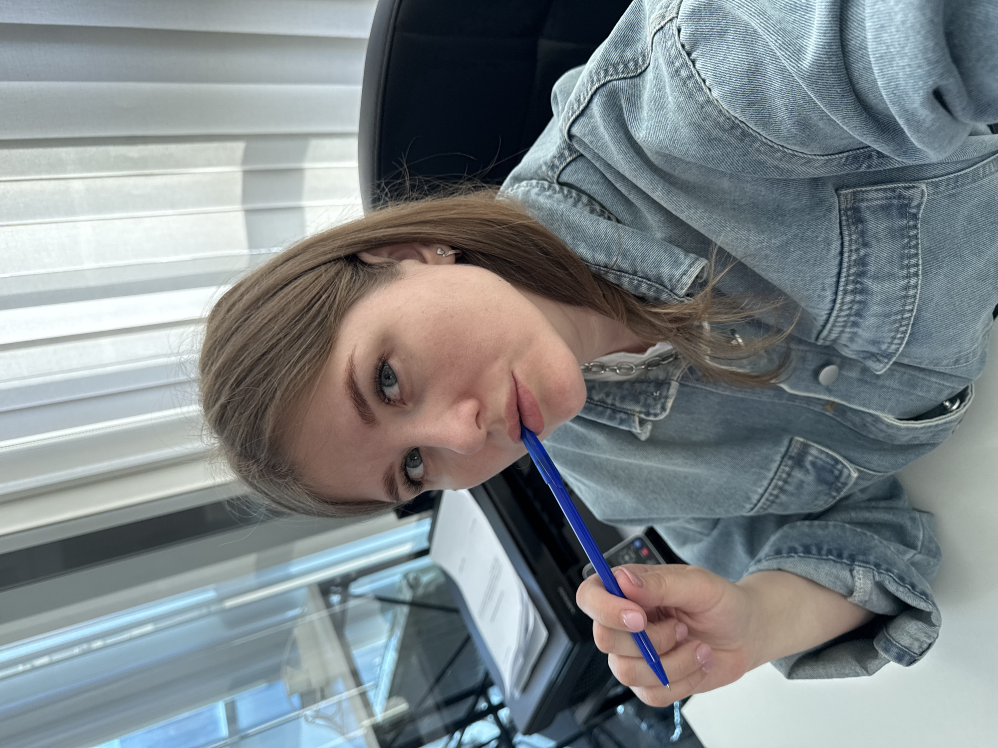 A young woman with light brown hair and blue eyes is seated in an office chair in what appears to be an office setting, with a white desk in front of her and venetian blinds visible in the background. She is wearing a light blue denim jacket. She is holding a blue pen to her lips. A printer sits on the desk next to her left hand, and a sheet of paper appears to be in the printer. The location is Perm, Russia.