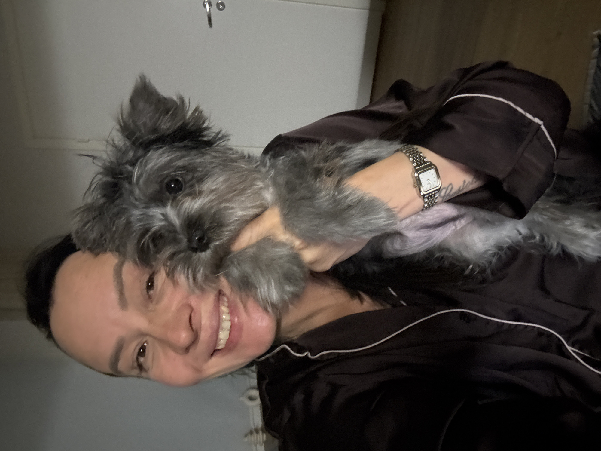 A woman with light skin, dark hair pulled back with a headband, and a broad smile holds a small, gray, furry dog close to her face. The dog's face is positioned near the woman's cheek, and she appears to be hugging it. The woman is wearing a dark-colored silky pajama-style top with white piping. On her left wrist, she wears a silver watch with a rectangular face. In the background, a white door is visible, along with a hint of another piece of furniture. The location in the city of Chaikovsky, Russia cannot be determined from the image.
