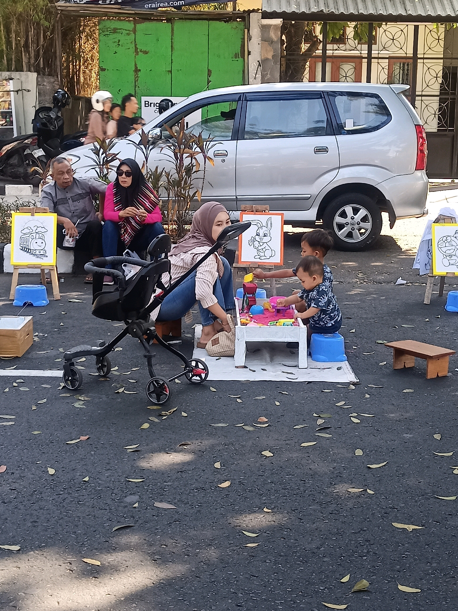 In Surakarta, Indonesia, a scene unfolds with a group of people gathered outdoors on an asphalt surface. A silver minivan is parked nearby, partially obscured by a woman in a hijab sitting on the ground. She is looking to her right, and her left hand rests on a white table laden with colorful toys. Two young boys are engaged with the toys on the table. One boy, wearing a dark shirt with a white pattern, is actively playing, while the other, in a blue shirt with a similar white pattern, is also interacting with the toys. A black stroller is positioned in front of the woman, and a wooden stool is located to the right of the playing area. In the background, a man in a gray shirt and a woman in a pink headscarf are seated, and a motorcycle with two helmets is visible. Further back, a green wall and a building with a metal fence can be seen. The ground is scattered with fallen leaves.