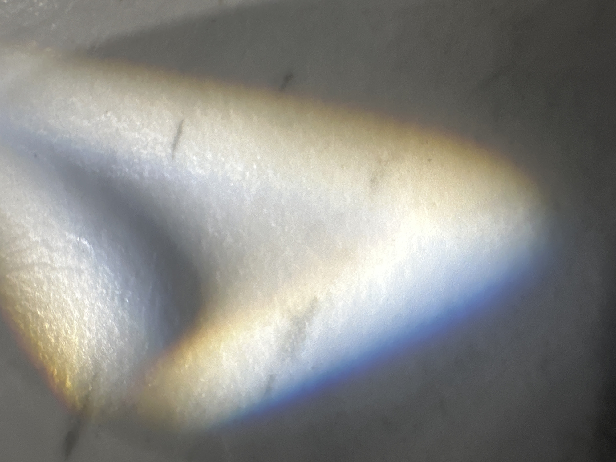 This media file is an abstract image that appears to capture the effect of light refracting through a prism or water. The primary subjects are bands of color, resembling a spectrum, that stretch across a textured, uneven surface. There are no visible people, recognizable objects, or clear scene or setting details.
The activity taking place is the interaction of light with a surface, creating a prismatic effect. Notable details include the presence of colors that suggest a rainbow or a similar optical phenomenon. The colors present are yellows, oranges, blues, and purples, blending into each other. The background surface is predominantly white or light grey, with subtle textures and what appear to be small dark specks, possibly dust or imperfections on the surface. The lighting conditions are not clearly discernible beyond the fact that light is present and interacting with the surface in a way that causes refraction. There are no location cues or emotional undertones that can be identified from the image itself. No text is visible in the media file.
