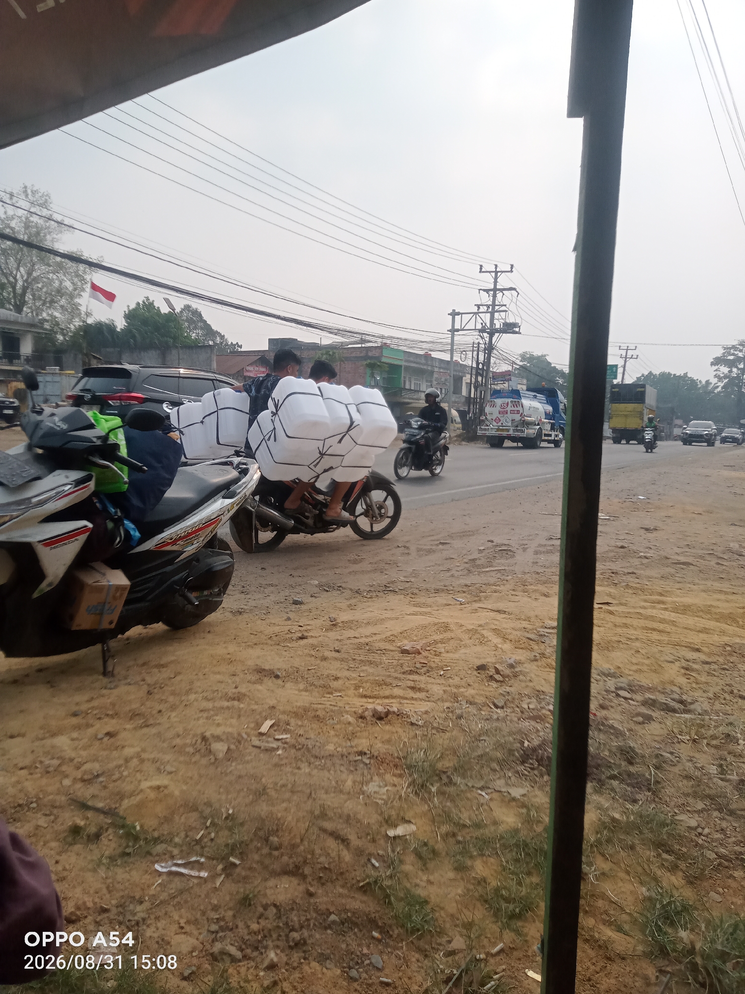 Two motorcycles are driving on a dirt road in Jambi, Indonesia. The motorcycle in the foreground is white and has a box strapped to the front. The motorcycle in the background is black and is carrying a large load of white packages. A man in a helmet is riding the black motorcycle, and two men are sitting behind him on the motorcycle. The men are wearing blue shirts and are carrying white packages. The road is dusty and there are trees and buildings in the background.