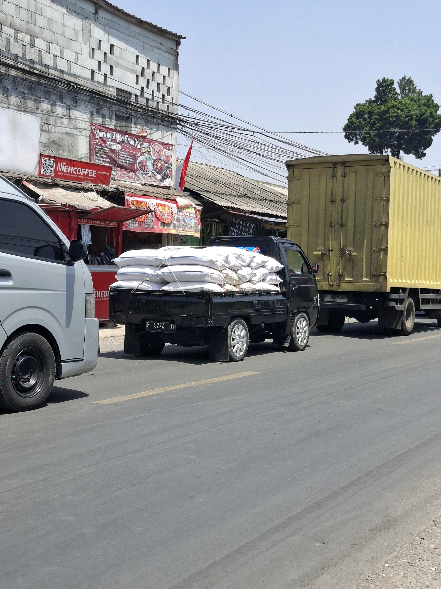 The image captures a scene on a paved road in Sukalarang, Indonesia, under a clear blue sky. In the foreground, a gray van is parked on the left side of the frame, with its front wheel and part of its body visible. To its right, a black pickup truck is stopped, its bed loaded with multiple white sacks secured with ropes. The truck's license plate reads "F 8224 UY". Behind the pickup truck, a large yellow shipping container truck is also stopped, with its rear wheels and container visible.
In the background, a two-story building with a rough, textured facade stands. Several signs are affixed to the building's front. One banner, with red and white colors and text in Indonesian, advertises "Warung Jajan Enak Ko" and displays images of food. Another sign, "NIEH COFFEE," features a QR code. Numerous electrical wires crisscross above the building and the street. A lone tree with dense green foliage stands in the distance, behind the yellow container truck. The road appears to have a yellow lane marker. The overall impression is a snapshot of a moment on a relatively busy street in Sukalarang, likely during daytime.