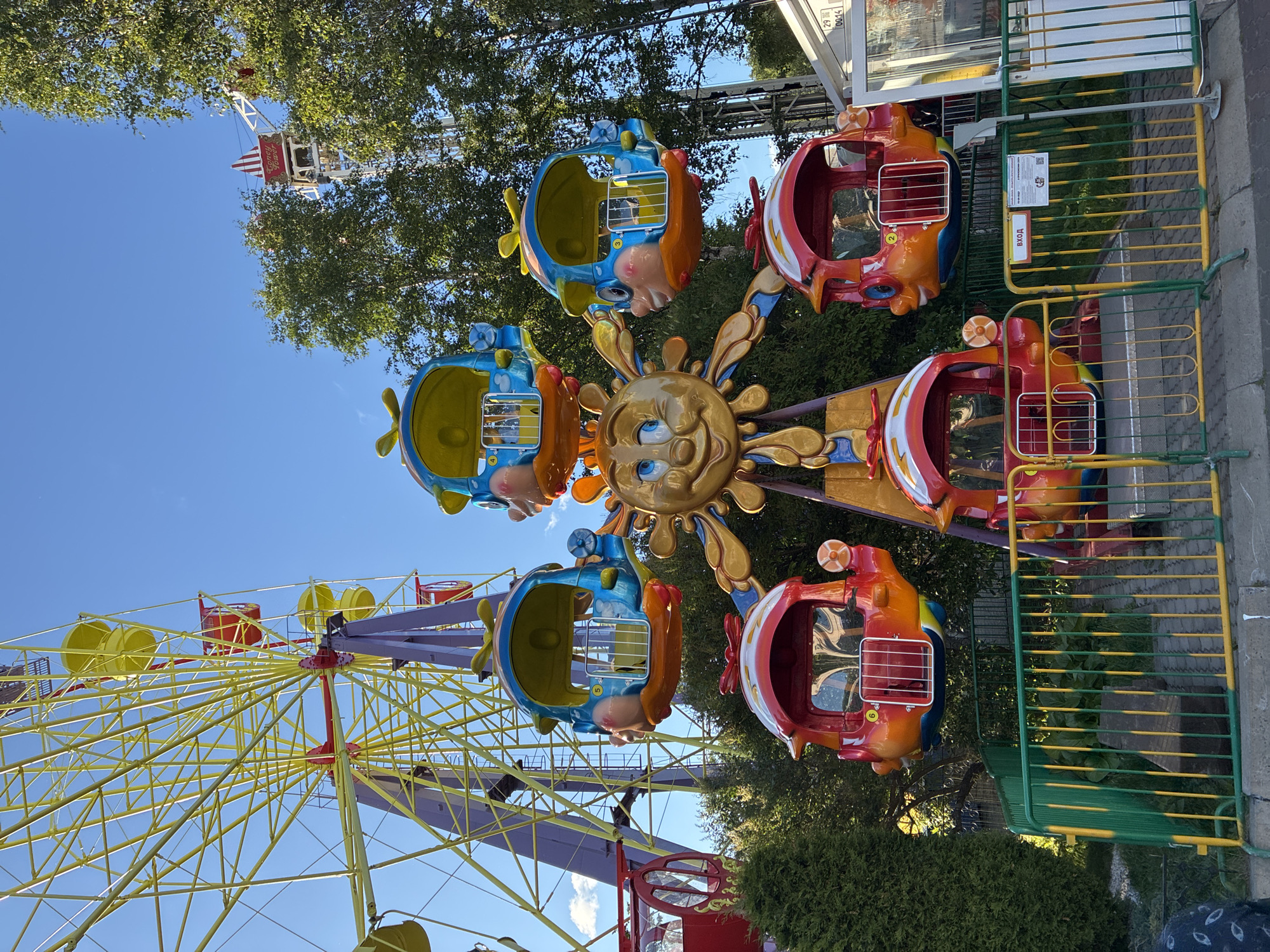 In a sunlit amusement park, a children's Ferris wheel features a central sun character with a smiling face. Attached to the sun's golden spokes are whimsical gondolas shaped like cartoon helicopters. Three of these gondolas are blue with yellow propellers and round, cartoonish eyes, while two are red and orange with white-capped tops and similar cartoonish features. The Ferris wheel's large yellow structure, with its intricate metal framework and purple support beams, is partially visible to the left. A bright blue sky serves as the backdrop, with lush green trees framing the scene on the right and scattered clouds. In the foreground, a yellow and green fence separates the ride area from a paved walkway and concrete border. The overall atmosphere is cheerful and playful, indicative of a family-friendly attraction.