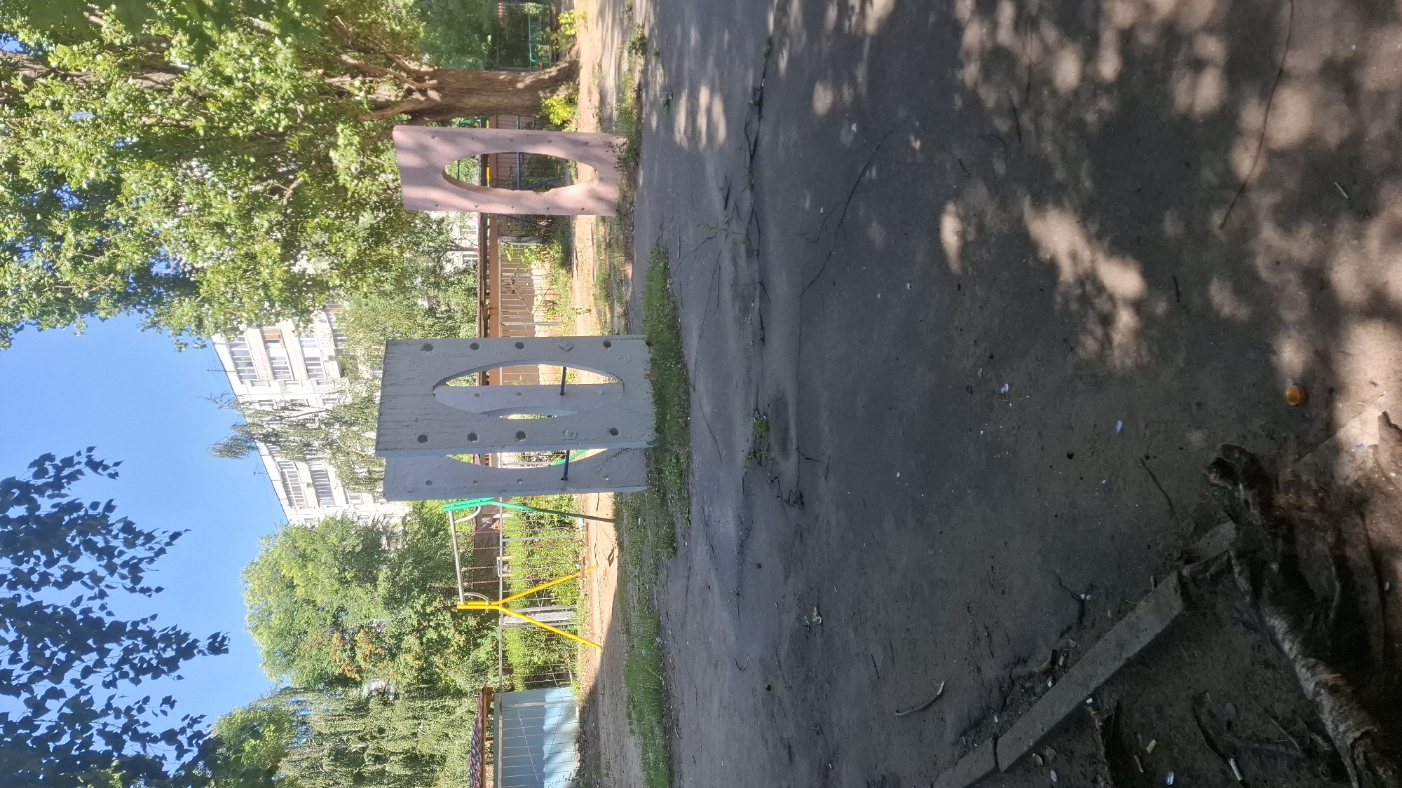 An outdoor, daytime scene features a playground in front of a residential building in Voronezh, Russia. The playground area consists of a concrete surface with cracks and patches of grass growing through. In the mid-ground, two abstract concrete structures with oval openings are present. The structure on the left is light gray with several circular indentations, and the one on the right is pinkish-brown. Behind these structures, a yellow metal swing set with green accents is visible. A fence, possibly chain-link, runs along the back of the playground. Beyond the playground, trees with green foliage are scattered, and a multi-story residential building with many windows stands in the background. The sky is clear and blue. In the foreground, a dark, rough surface is visible, with some tree roots or branches lying on it, along with a small orange object that may be a plastic cap. Shadows of leaves are cast across the ground, indicating sunlight filtering through trees. No people are visible in the image.