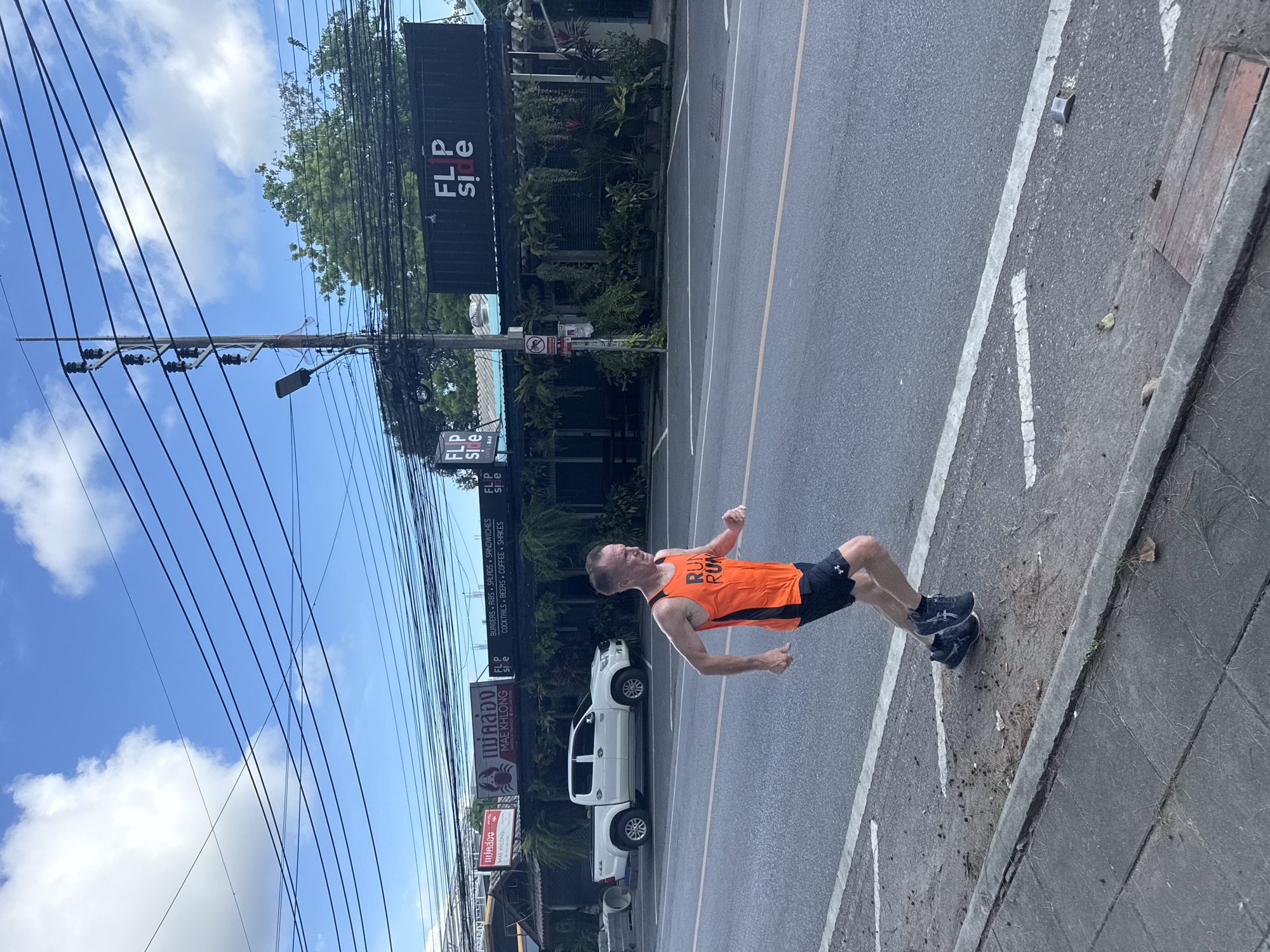 A Caucasian male runner is seen from the chest down, wearing an orange tank top with "RUN RUN" printed on it and black shorts. He is mid-stride on the right side of a paved road, with his body angled towards the left of the frame. The road has a white dashed line separating the lanes and a solid white line marking the shoulder. In the background, a white pickup truck is parked on the left side of the road, behind a concrete barrier. Further back, a building with a dark facade and hanging plants is visible, identified by signs as "FLIP SIDE" and "MAE KHLONG." The sky above is blue with scattered white clouds, and a complex network of electrical wires is strung across the frame. The scene appears to be in Rawai, Thailand, given the presence of Thai script on signage and the general aesthetic.