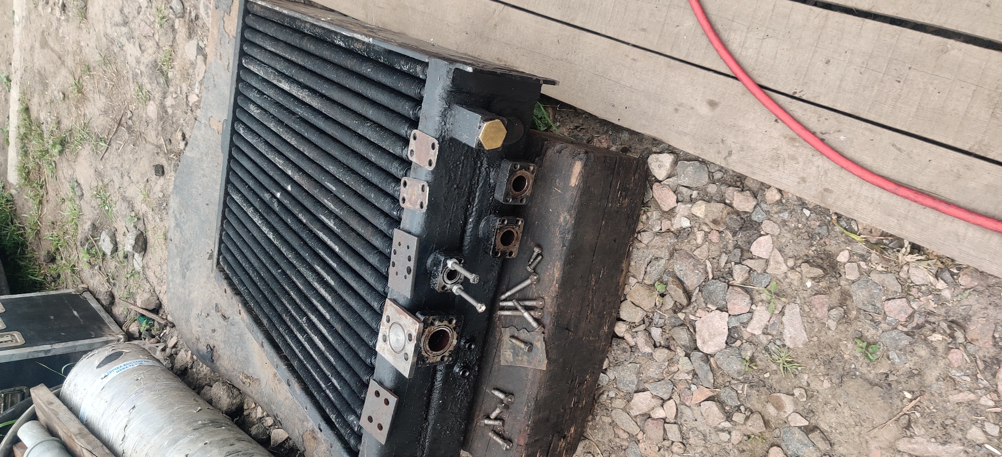 The image shows a close-up, high-angle view of a black radiator with a gridded surface, likely for an industrial or automotive application, lying on a wooden surface and next to a pile of gravel. Several metal plates and bolts are scattered around the radiator. To the left of the radiator, partially visible, is a large, cylindrical metallic object with blue text. In the background, a black case and some vegetation are visible. A red hose is visible on the wooden surface in the lower right corner. The setting appears to be an outdoor area, possibly a workshop or storage yard. The image does not contain any people or identifiable landmarks that would allow for a specific location within Rizunenkove, Ukraine.