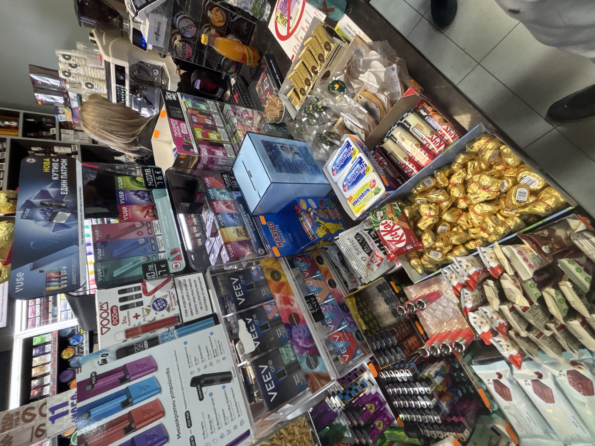The image captures a cluttered display at a retail location in Zlatograd, Bulgaria, possibly a small grocery or convenience store. The display showcases a variety of products including disposable vapes (VUSE, VOOM, VEEV), cigarettes, and confectionery items such as KitKat, Kinder, and Haribo. A person with blonde hair is partially visible near the cash register. The items are densely packed, and prices are displayed in both Bulgarian Lev (лв) and Euros (€). The store has tiled flooring and appears to be located in a commercial area.