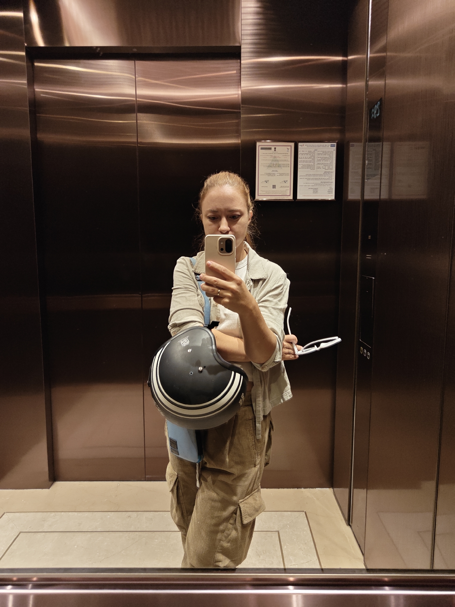 A woman is taking a selfie in an elevator. She is wearing a light-colored shirt, cargo pants, and is holding a motorcycle helmet and glasses. The elevator is modern with bronze-colored metal walls. Two certificates are affixed to the back wall. The floor of the elevator is light-colored and tiled. Based on the certificate on the back wall written in Hindi and the general appearance of the elevator and surroundings, this is likely located in the city of Mumbai, India.