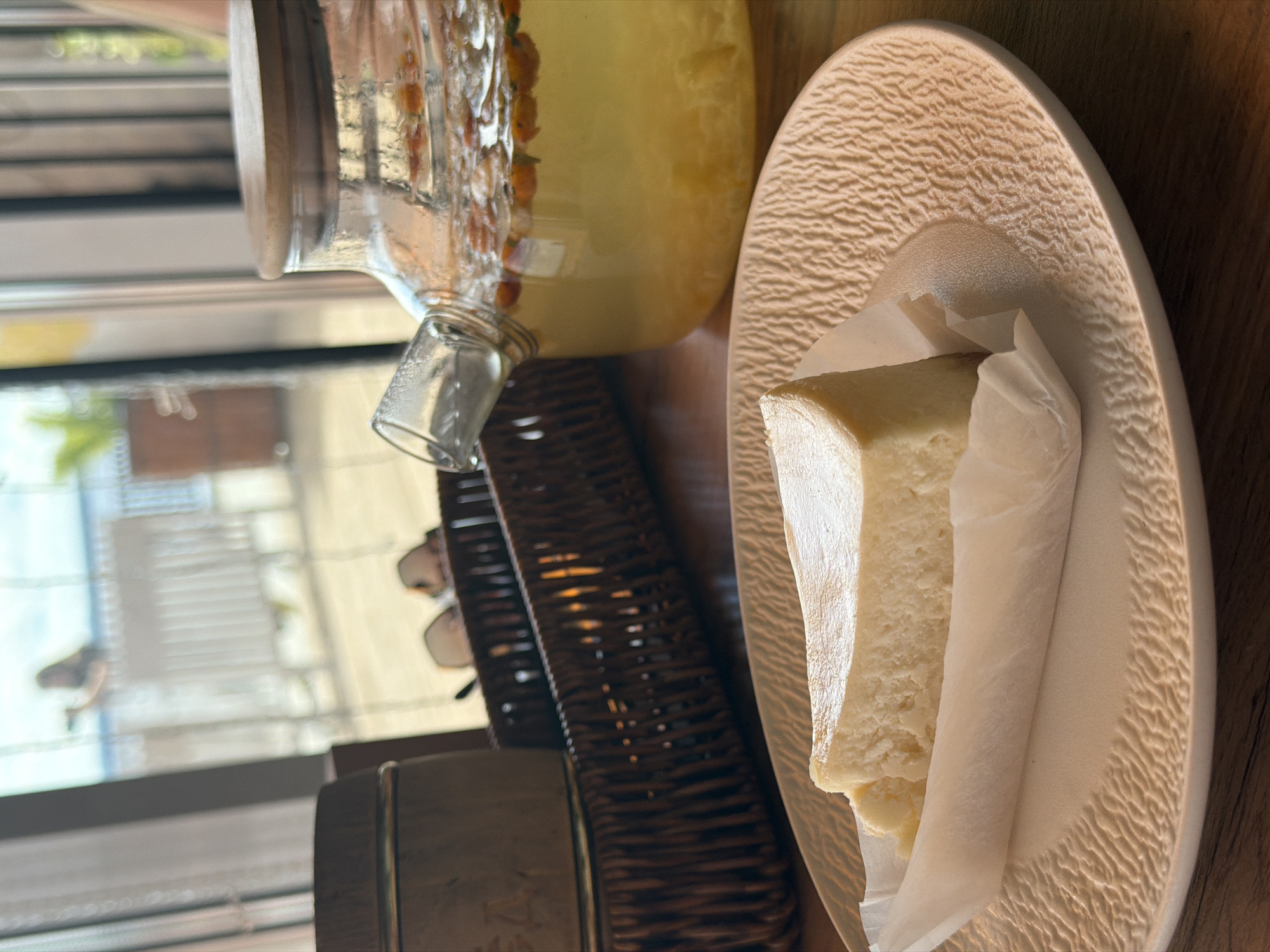 A slice of cheesecake sits on a textured, cream-colored plate. The cheesecake is triangular and wrapped in white parchment paper. To the right of the plate, a glass teapot filled with a yellow beverage and orange berries is positioned on a dark woven tray. In the background, a blurry figure stands near a white railing, possibly overlooking a body of water. The scene appears to be set indoors, with a large window providing a view of the outdoors. The location is identified as Chaikovsky, Russia.
