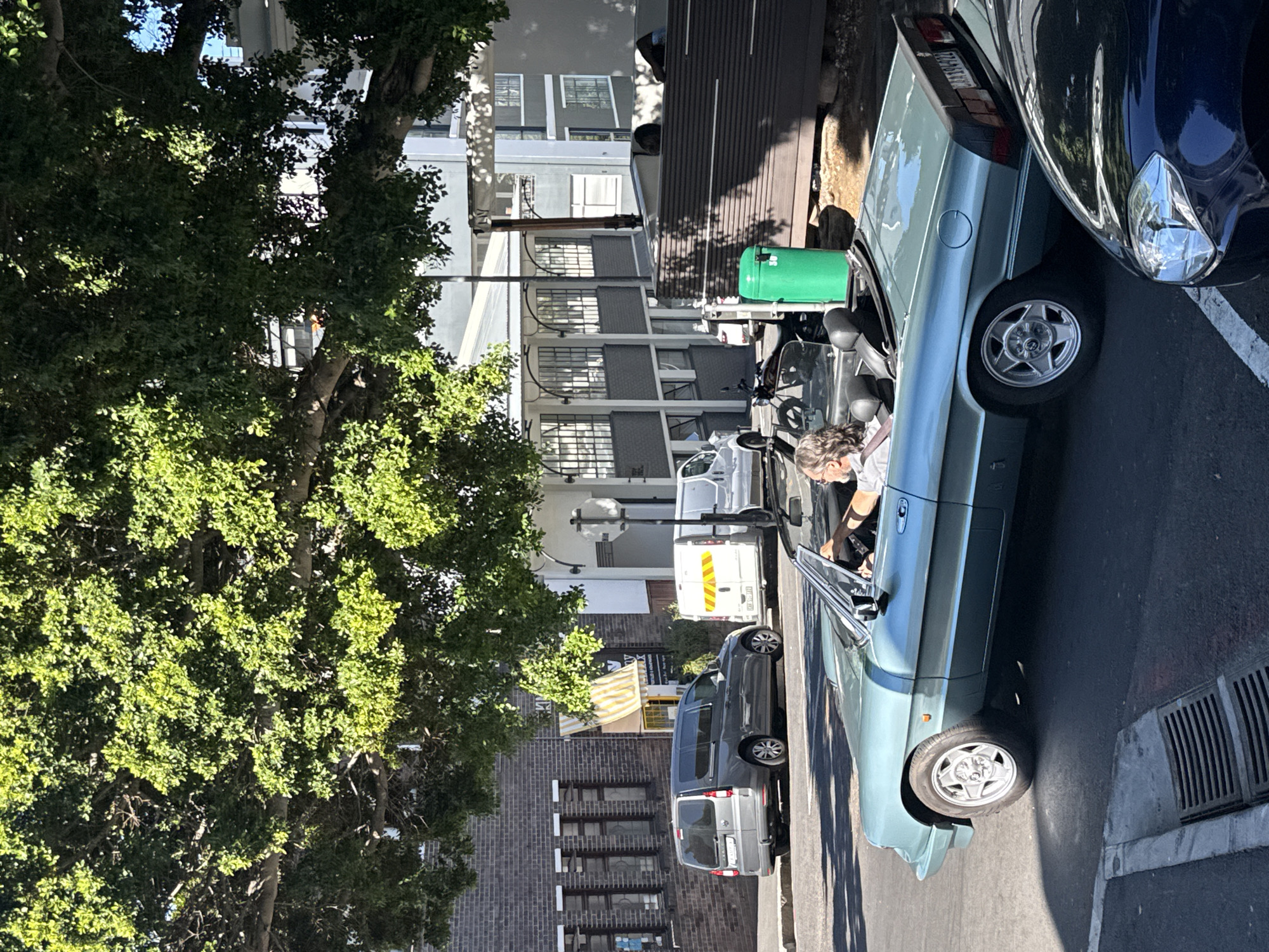 A street scene in Cape Town, South Africa, shows a light blue convertible parked on the right side of the street next to a black vehicle. An individual with gray hair is sitting in the driver's seat of the convertible. A green trash bin is behind the convertible. Several vans and a small truck are parked further down the street. Buildings with varying architectural styles are visible on both sides of the street, including brick buildings and white buildings with large windows. A large tree with dense green foliage is on the left side of the street. The street appears to be in a residential or commercial area, with a mix of vehicle traffic and pedestrian activity.