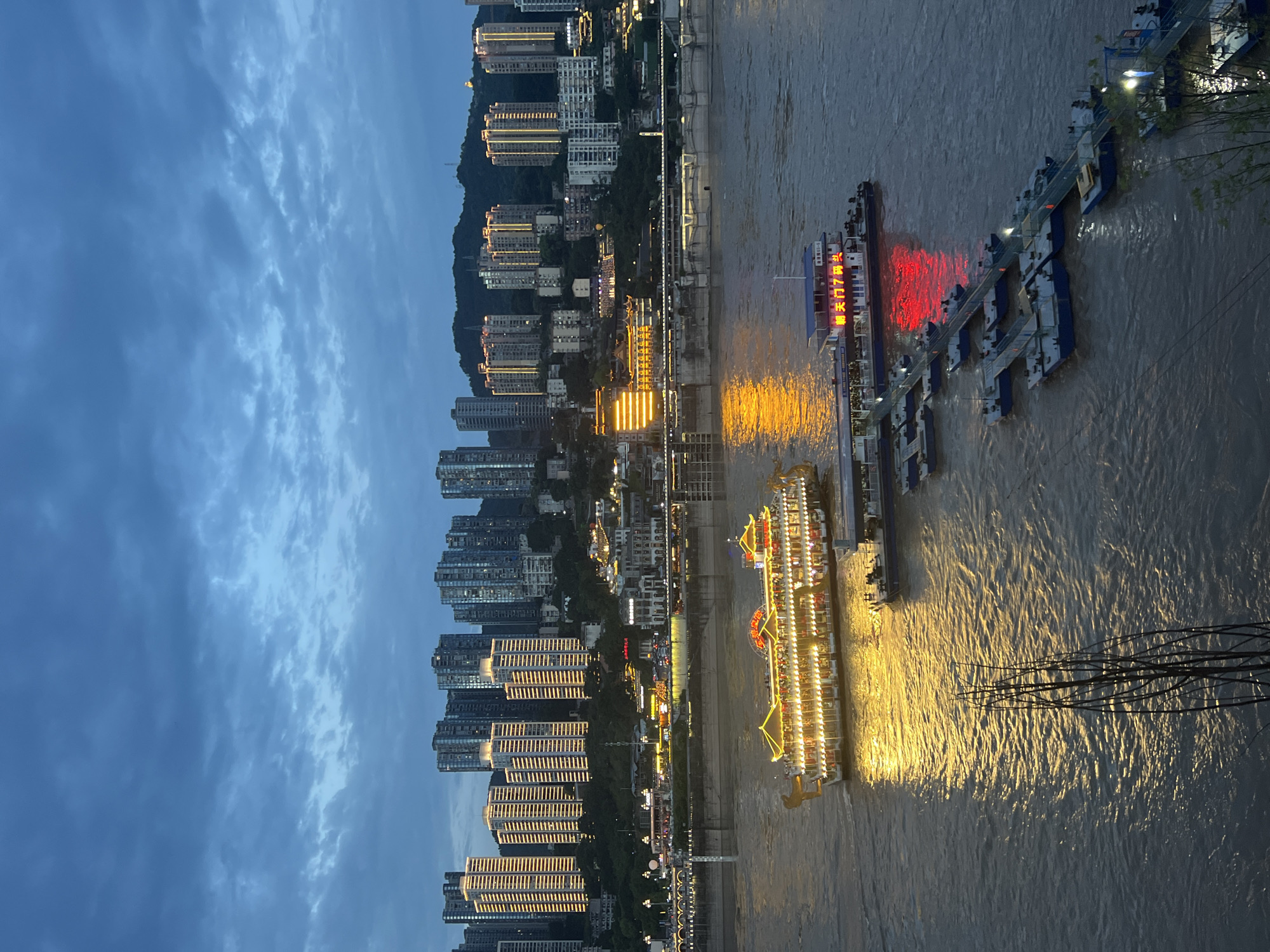 This is a wide, high-angle, outdoor, medium shot of the Yangtze River at dusk, featuring the Yuzhong District, Chongqing, China. In the foreground, the murky brown water of the river is visible, with reflections of lights from the surrounding cityscape. A large, illuminated boat with a distinctive golden roof and ornate dragon-like prows is sailing on the river. Several smaller, flat-bottomed boats are moored nearby. In the middle ground, a riverfront promenade is visible, lined with buildings that are lit up. The background is dominated by a hillside covered with dense trees, and towering over the trees are numerous modern high-rise apartment buildings and skyscrapers, many of which are illuminated with warm-toned lights. The sky is a deep blue, with scattered, textured clouds. The overall scene conveys a vibrant and bustling urban environment at twilight.