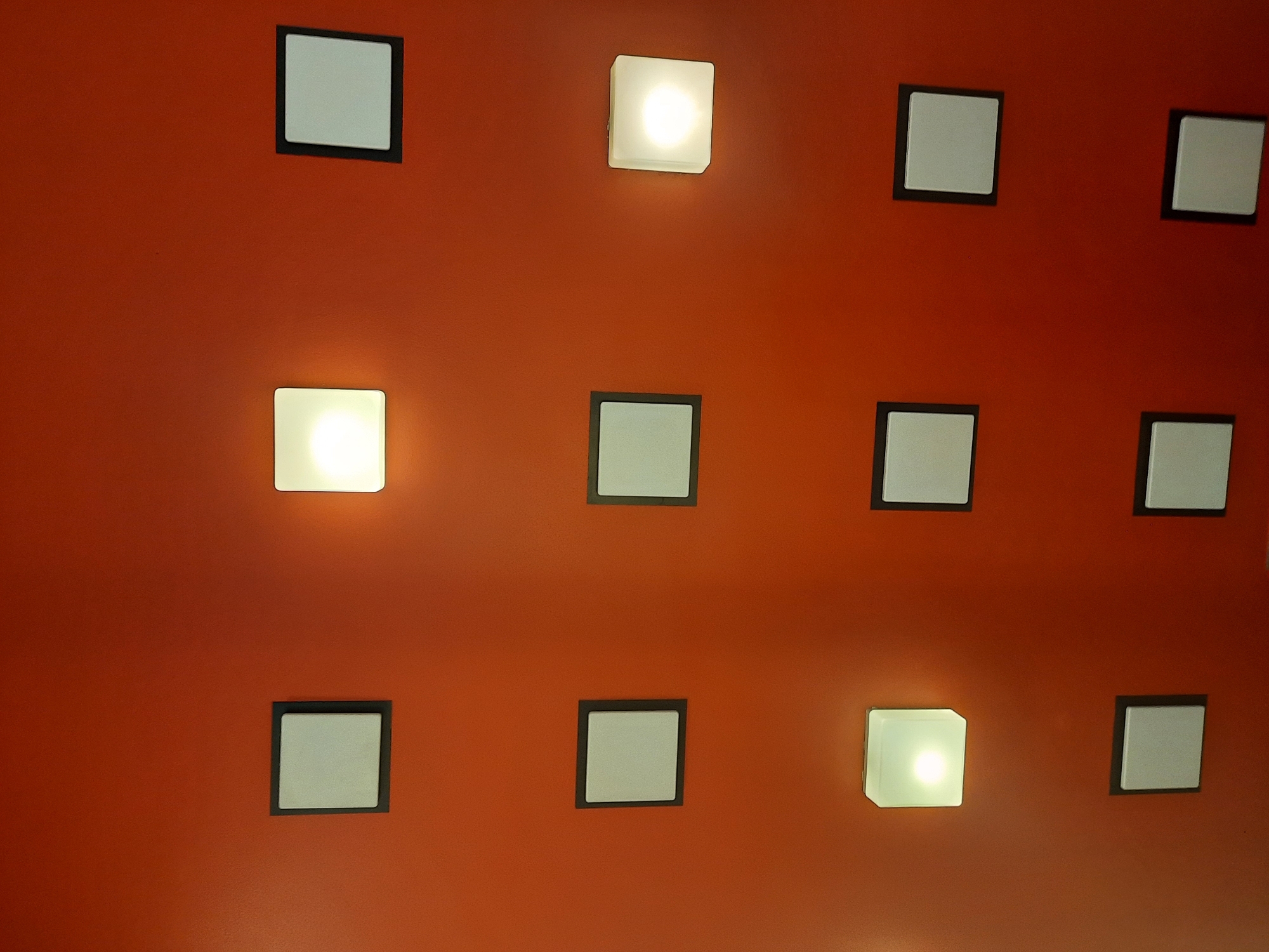 The image displays a wall with a grid of square lights. There are three rows of three lights each, totaling nine lights. Some of the lights are illuminated, emitting a warm white glow, while others appear to be unlit. The wall is painted a uniform reddish-orange color. The lights are mounted flush with the wall and have a dark border around a central square panel. The scene appears to be indoors, possibly in a hallway or public space, given the organized arrangement of the lights. No people or other objects are visible in the frame.