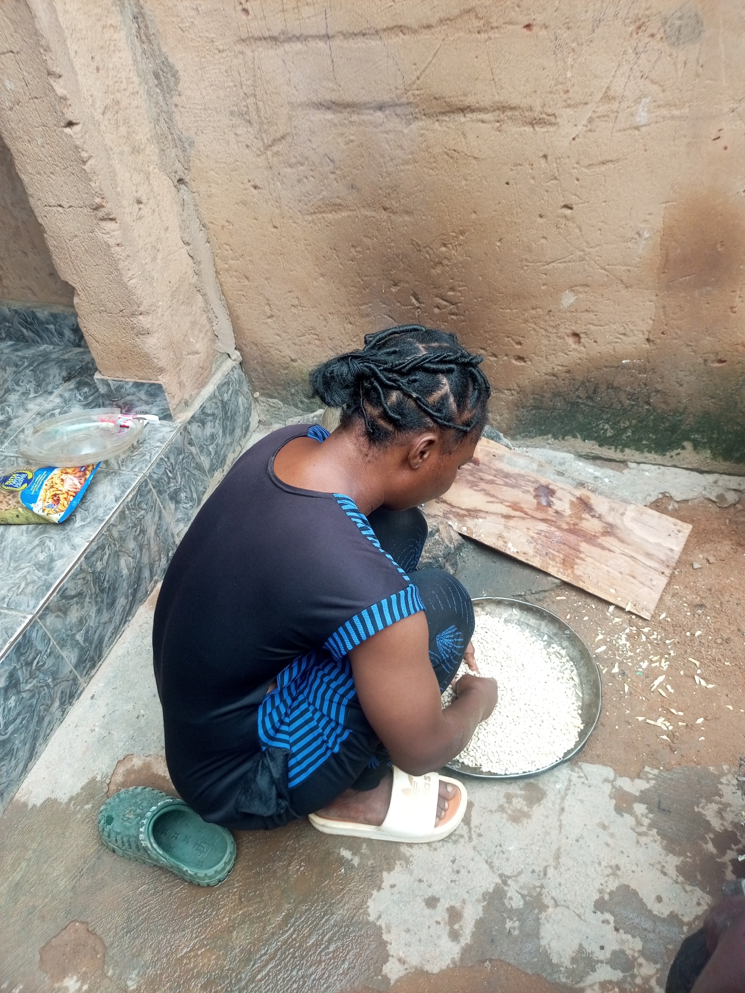 A woman with braided hair is kneeling on the ground in what appears to be a courtyard or outdoor preparation area. She is wearing a black t-shirt with blue stripes on the sleeves and black pants with blue accents. She is sorting through a large quantity of small white grains in a metal tray with her hands. To her left, on a set of tiled steps, are a bag of pasta and a clear plastic bowl. To her right, a piece of weathered wood lies on the ground. The ground is made of concrete and dirt, with some puddles of water present, suggesting it may have recently rained. The background is a rough, textured wall. A green rubber sandal is visible on the ground to her left, and a white sandal is on her right foot. The scene is likely in a residential area of Gombe, Nigeria, given the style of the building materials and the presence of common household items.