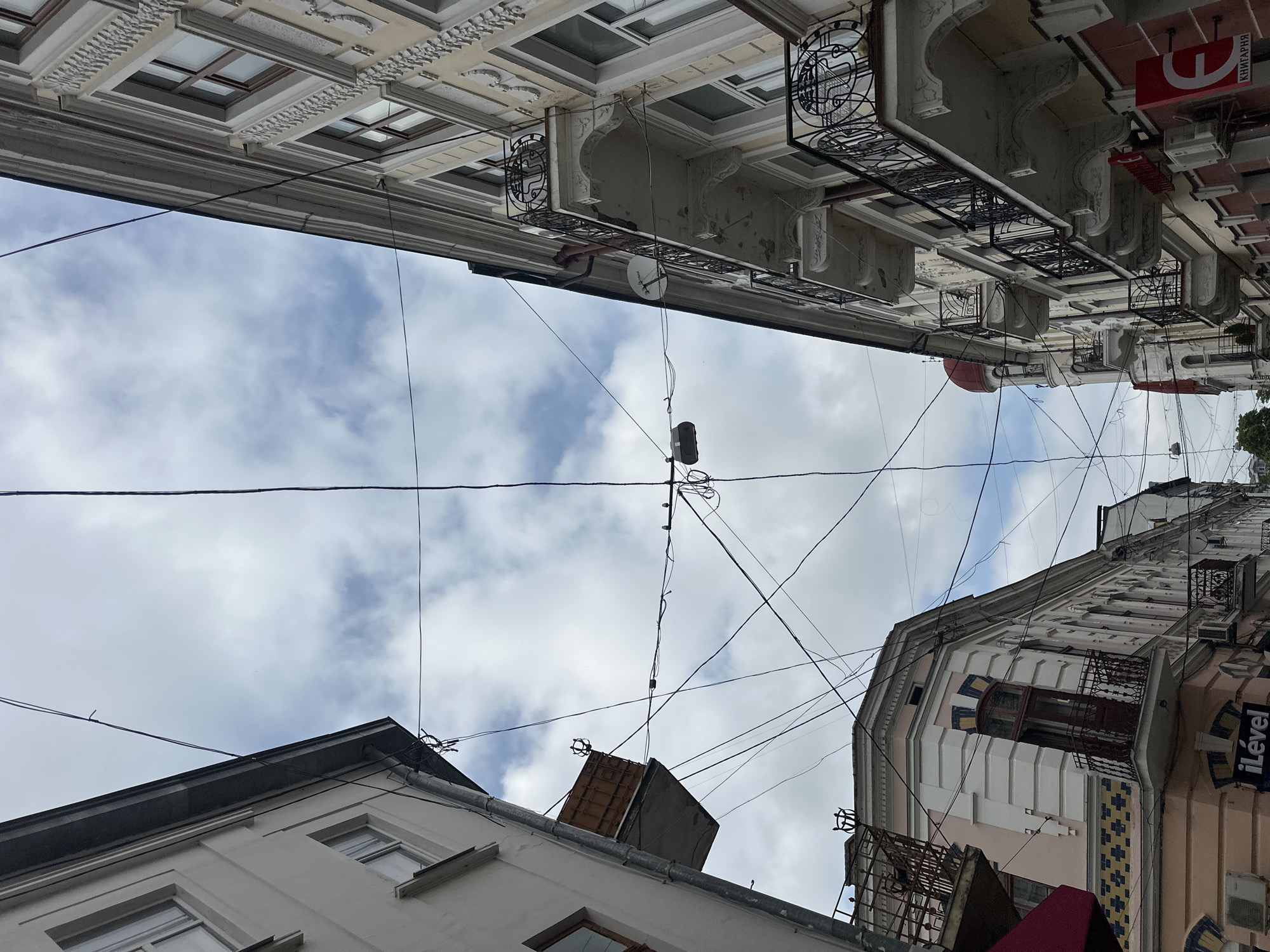 A vertical perspective looking upwards captures a narrow section of the sky framed by multi-story buildings with historical architecture in Ternopil, Ukraine. The sky is predominantly overcast with scattered blue patches. A dense network of black electrical and communication wires crisscrosses the sky, centrally suspending a single street light fixture.
On the left side of the frame, a multi-story building with a plain white facade is visible, featuring simple windows, a prominent gray downspout, and a distinctive enclosed rectangular balcony structure finished in a rust-brown color. Below this, a pink-hued corner building displays decorative blue and yellow tiled patterns, an arched window with an ornate wrought-iron balcony, and a ground-level sign reading "iLével" in white text on blue. Air conditioning units are affixed to both the white and pink buildings.
On the right side of the frame, a cream-colored building showcases intricate stucco ornamentation, featuring multiple stories with ornate wrought-iron balconies. A white satellite dish is mounted on one of these balconies. At the lower right, a red storefront sign displays a stylized epsilon-like character and the Ukrainian word "КНИГАРНЯ" (bookstore). In the background on the right, a building with a prominent red domed roof is partially visible, with another smaller red dome behind it.
The scene is devoid of human presence.