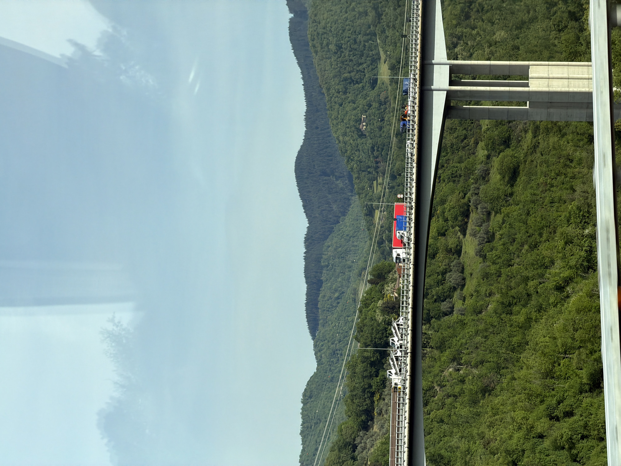 The image shows a high-altitude view, most likely from the new Variante di Valico road. The location is identifiable as Barberino di Mugello, Italy. The scene features a concrete bridge structure on the right side of the frame, with a dense green forested area underneath it.
On the left side, a landscape of rolling mountains covered in dense trees is visible, stretching into the distance. The sky above is a light blue with faint cloud cover.
A series of vehicles, including a red-and-blue truck, are visible traveling across the bridge structure along the edge of the frame. These vehicles are the sole indication of human activity within the scene.