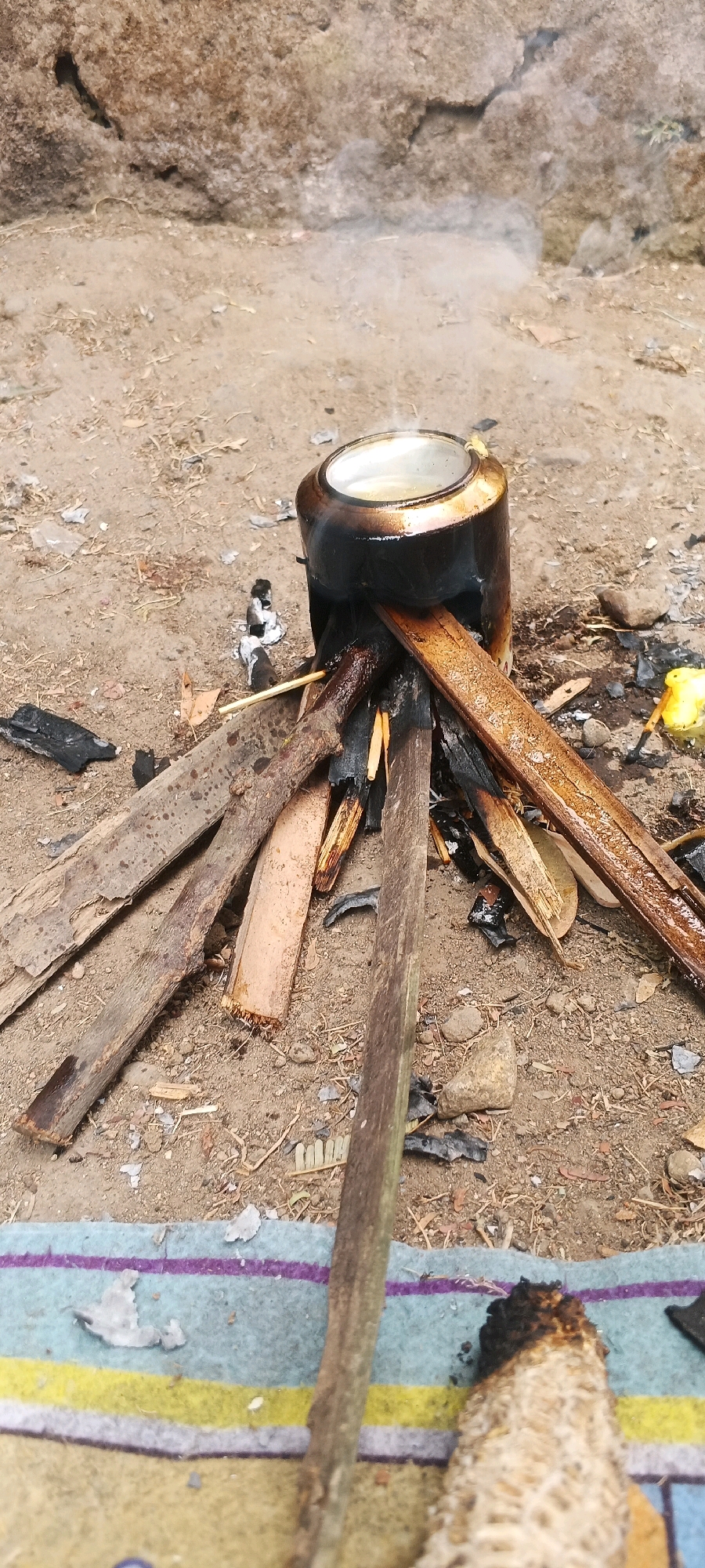 A close-up, ground-level shot captures an outdoor scene at an unspecified location within Penanggungan, Indonesia, where a pot of liquid is being heated over a small, makeshift fire. The pot, dark and cylindrical with a metal lid, rests on a base constructed from several pieces of firewood, some of which are charred. Wisps of steam rise from the pot, indicating the liquid is hot. The fire itself consists of burning embers and wood fragments scattered on the dry, dirt ground. The surrounding environment appears rustic, with dirt and small stones forming the ground surface, and a textured, rocky incline visible in the background. A colorful textile, striped with blue, purple, and yellow, is laid out in the foreground, partially covered by a burnt corn cob and a wooden stick. Debris, including bits of paper and charcoal, is scattered around the fire pit, suggesting a utilitarian or informal cooking setup.