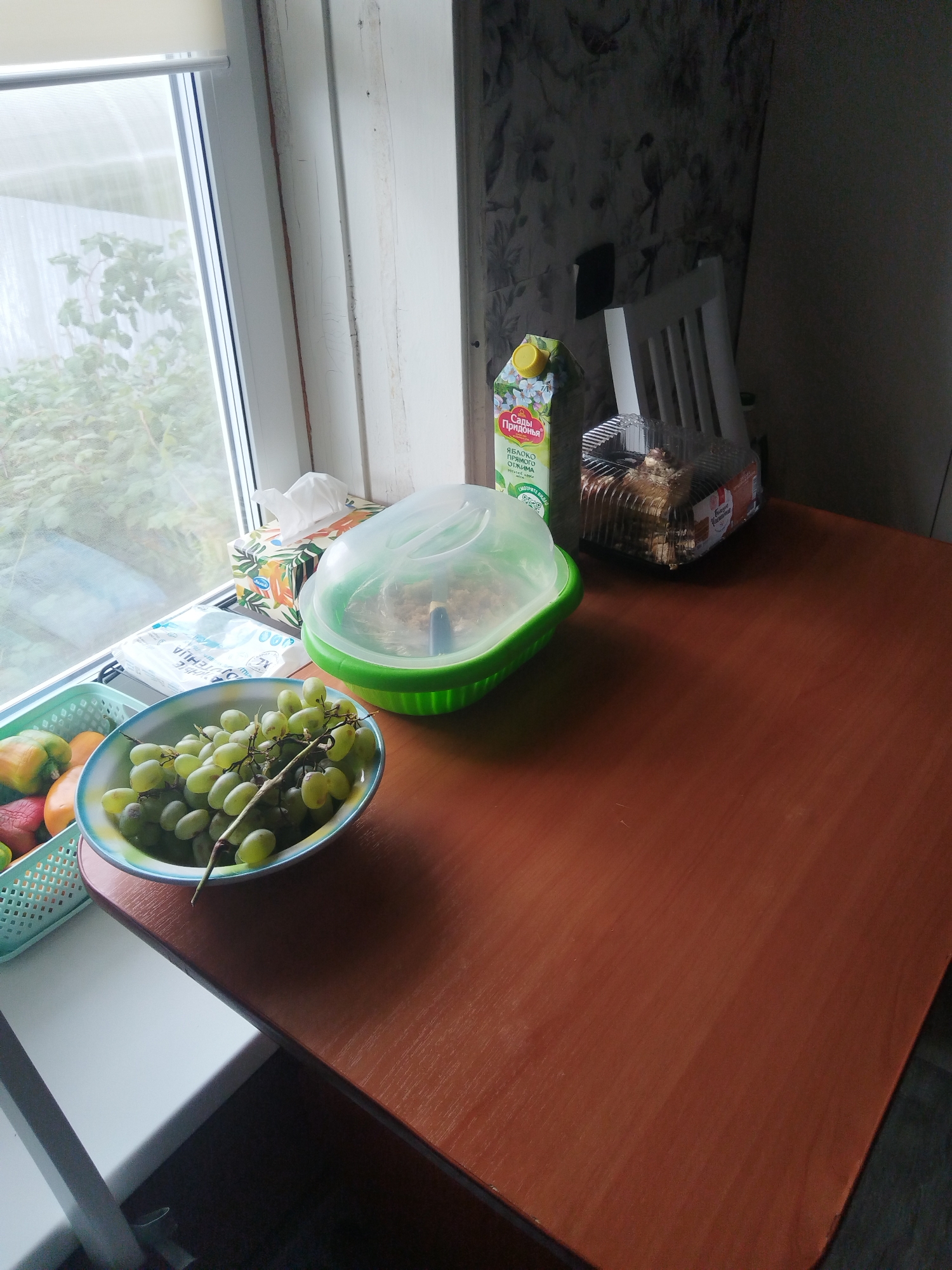 A still life scene unfolds on a wooden table near a window. In the foreground, a bowl of green grapes sits on the table, partially obscuring a light blue plastic basket containing various fruits, including what appears to be bell peppers and possibly oranges or peaches. To the left, a white tissue box with an orange and green floral pattern rests on the windowsill. Behind it, another white package with text is visible. A green plastic container with a clear lid, holding some food item, is situated on the table. Further back, a carton of apple juice with a yellow cap and a label featuring flowers stands next to a clear plastic container holding a cake or pastry. A white chair is positioned behind these items. The background shows a wall with a floral wallpaper pattern and a window with a white blind, revealing greenery outside. The scene appears to be set indoors in what could be a kitchen or dining area. There is no indication of the city of Votkinsk, Russia, within the image.