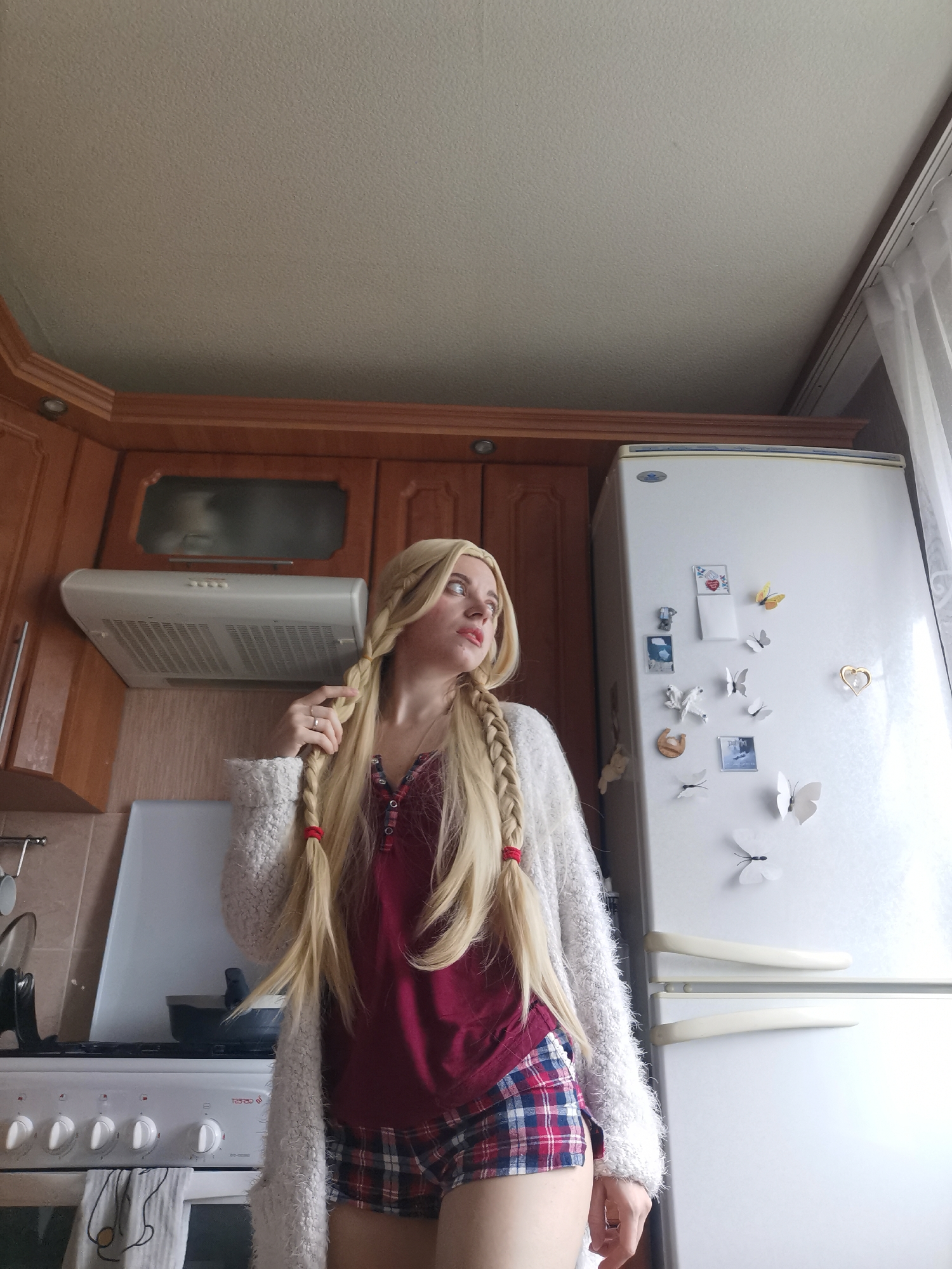 A blonde woman with long, braided hair stands in a kitchen, looking away from the camera. She is wearing a burgundy shirt and plaid shorts, with a fuzzy white cardigan. A white refrigerator covered in butterfly magnets stands to her right, and a white gas stove is to her left. Above the stove is a built-in range hood, and above that are wooden cabinets. The ceiling is a textured off-white.