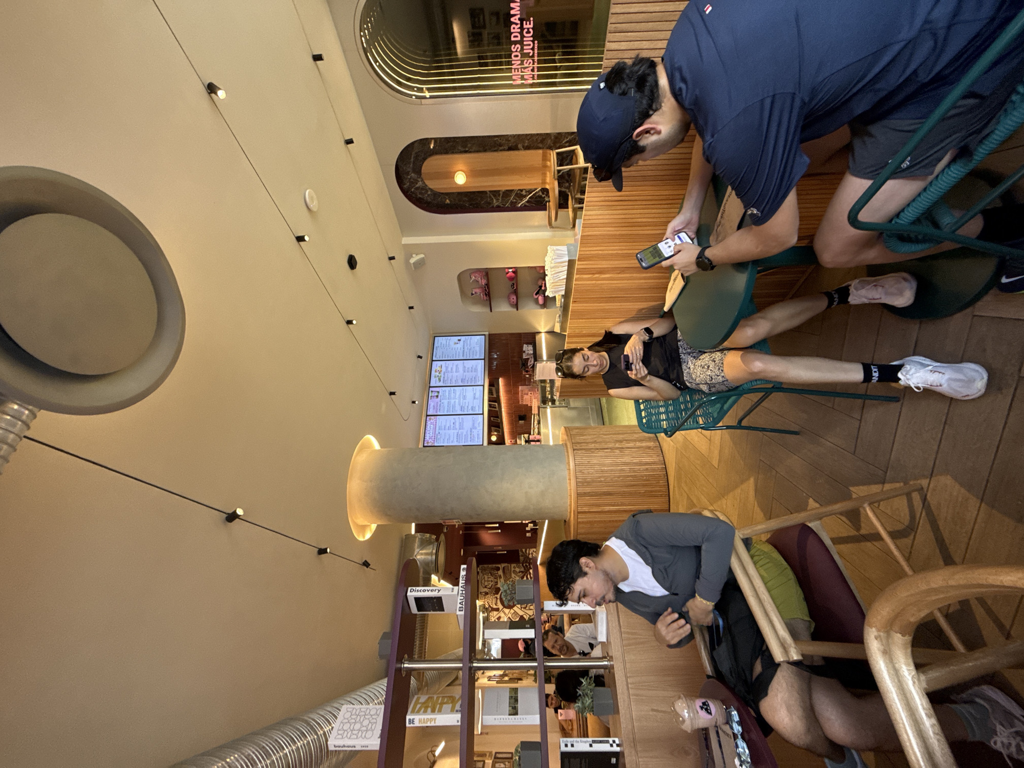 The image shows the interior of a café in Mexico City, Mexico. The café has a modern design with light-colored walls, wood paneling, and a variety of seating options. There are three people in the scene. Two men are sitting at a green table, and one is looking at a phone. The other man is sitting down and also looking at a phone. There is another man seated at a wooden table in the opposite direction of the other two. The environment has a bookshelf filled with various items and books. The lighting is warm, with several ceiling lights providing illumination. A menu board is visible in the background, suggesting a variety of food and drinks are available.