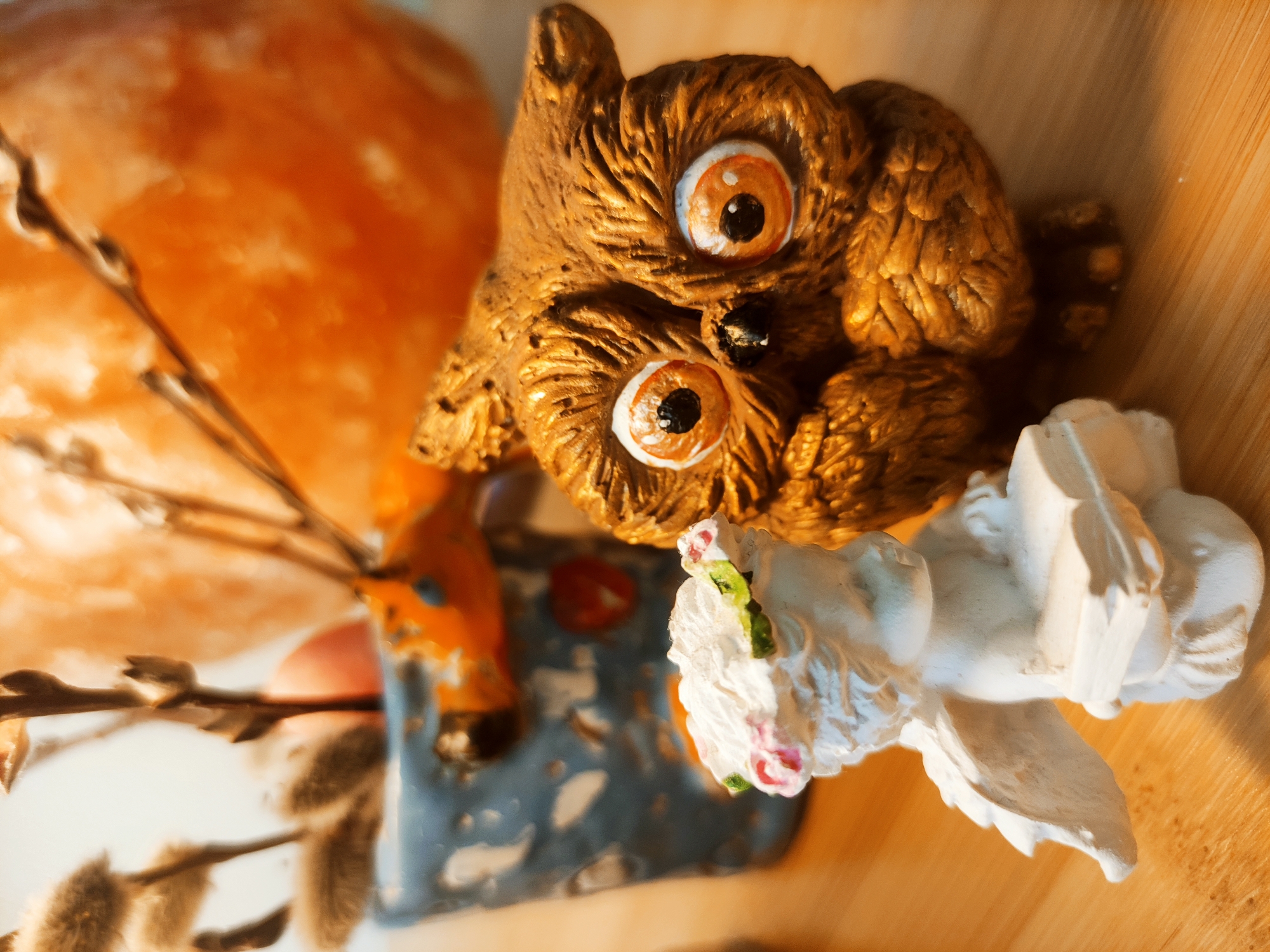 A close-up shot captures a still life scene of Easter decorations arranged on a light-colored surface, possibly wood. Dominating the composition is a brown, textured owl figurine with large, orange-rimmed eyes and a black beak. Next to the owl sits a white angel figurine with delicate features and floral accents. Behind the figures, a vase holds pussy willow branches, with an orange, egg-shaped object partially visible in the background. The light source creates strong shadows and highlights, emphasizing the textures and details of the decorations. The scene appears to be indoors, and the objects are arranged to create an Easter-themed display.