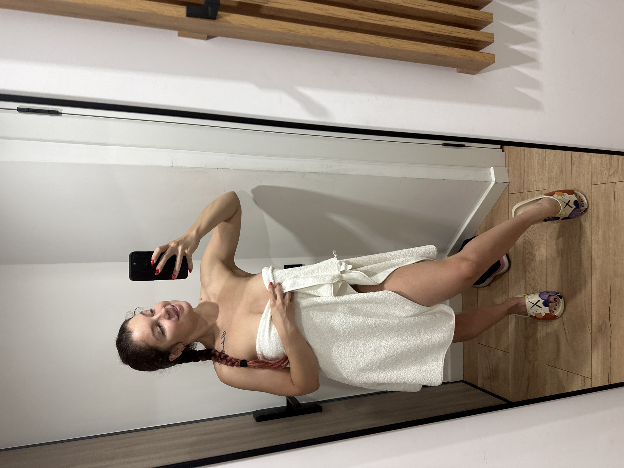A woman is taking a mirror selfie in what appears to be a fitting room or bathroom. She is wearing a white towel wrapped around her body and has her hair in a braid. She is holding a black smartphone in her right hand, and her left hand is holding the towel closed. She is wearing brightly colored slippers on her feet. The background shows a wooden floor, a white wall, and a closet door on the left. There is also a wooden structure on the right side of the image. The woman has a tattoo on her chest and her nails are painted red. There is no indication of the specific location being Moscow, Russia.