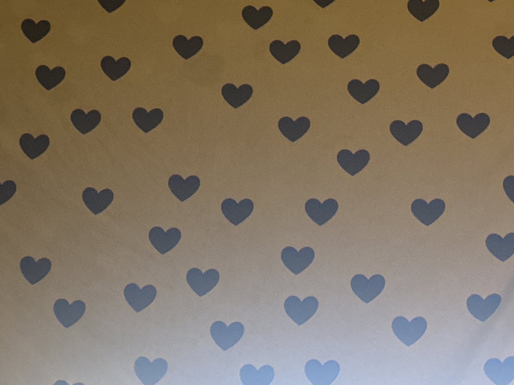 The image displays a close-up, top-down view of a brown, textured surface adorned with a repeating pattern of dark grey heart shapes. The hearts are uniformly sized and arranged in a staggered grid formation across the frame. The lighting suggests a somewhat dim environment, casting subtle shadows that enhance the three-dimensional appearance of the heart shapes. There is no indication of any specific location in Cheltenham, United Kingdom, or any people, actions, or interactions. The scene is purely a still life of a patterned surface.