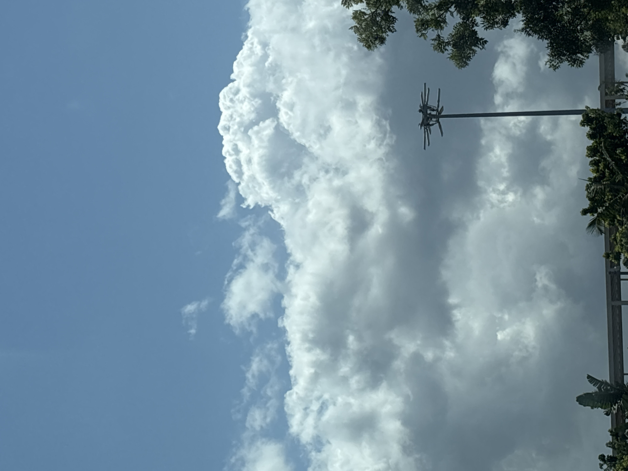 The image shows a view of the sky above a city, with a tall street light pole in the foreground. The sky is filled with a mix of cumulus and stratus clouds, with patches of blue visible. A large, fluffy cumulus cloud is prominent in the upper right portion of the image. The street light pole is tall and slender, with a multi-pronged light fixture at the top. Trees and a structure that appears to be part of an elevated walkway are visible at the bottom of the image. The scene is devoid of people and any specific identifying landmarks that would place it in Lisbon, Portugal.