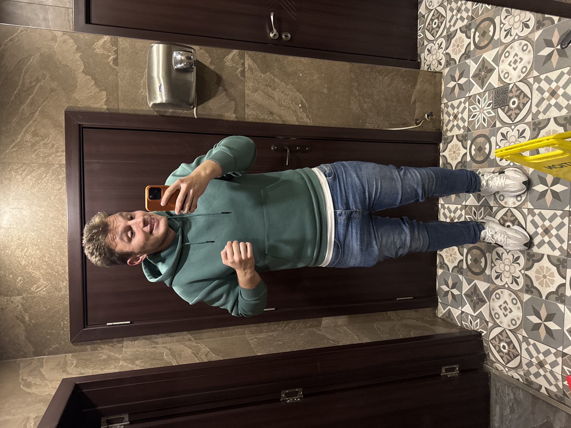 A full shot of a man standing in a tiled bathroom in Burgas, Bulgaria, likely a public restroom. He is holding an orange smartphone, presumably taking a selfie. He's wearing a light green hooded sweatshirt, blue jeans, and white sneakers. His expression is jovial. Behind him, there is a dark brown door with a silver hand dryer on the wall to the left. The floor is tiled with patterned, grey and beige tiles. A yellow caution sign is visible near his feet on the right. The walls are covered in rectangular tiles with a mottled, stone-like appearance. There is a second, darker door frame above the first.