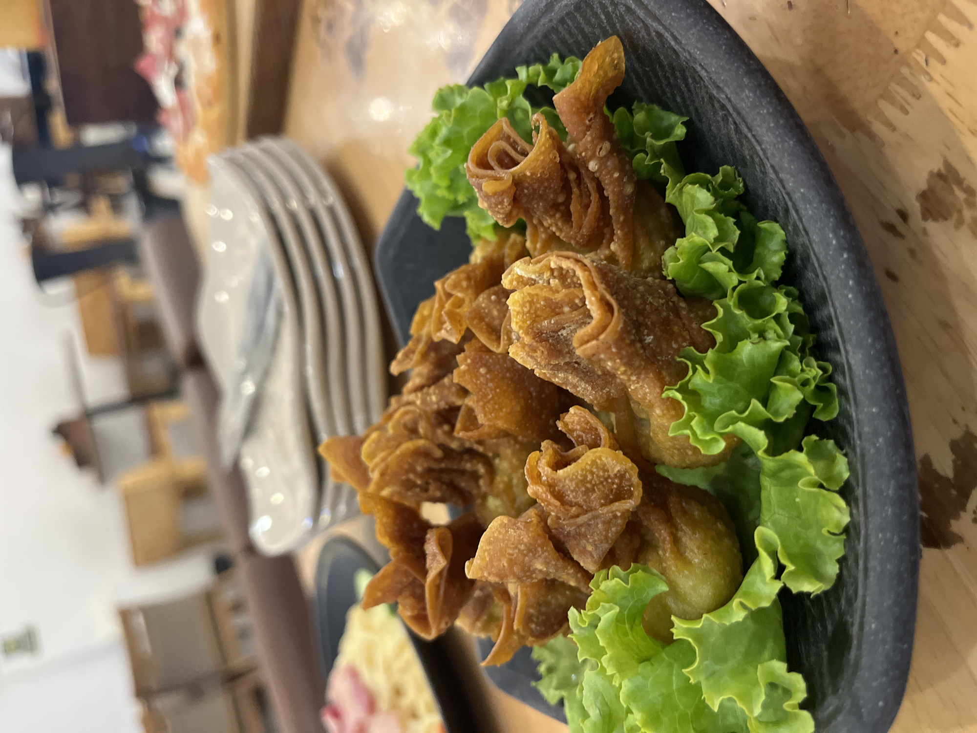 A close-up shot of a plate of fried wontons, likely crab rangoon, is arranged on a black plate and garnished with lettuce. The scene is indoors, possibly in a restaurant. A stack of plates and another dish with food are visible in the background. The location appears to be Bac Nha Trang Ward, Vietnam.