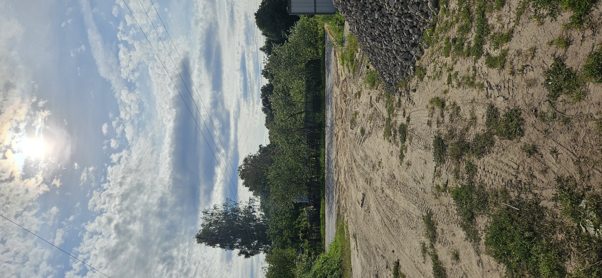 The image depicts an outdoor scene with a dirt foreground, a line of trees and a fence in the middle ground, and a bright, cloudy sky in the background. The dirt area shows tire tracks from a vehicle, suggesting recent activity. A pile of grey stones is visible on the right side of the foreground. The middle ground features a dark wooden fence bordering a lush green area with several trees, some of which appear to be fruit trees. Overhead, power lines crisscross the sky. The sun is visible through a break in the clouds, casting a bright light. The overall impression is of a rural or semi-rural setting.