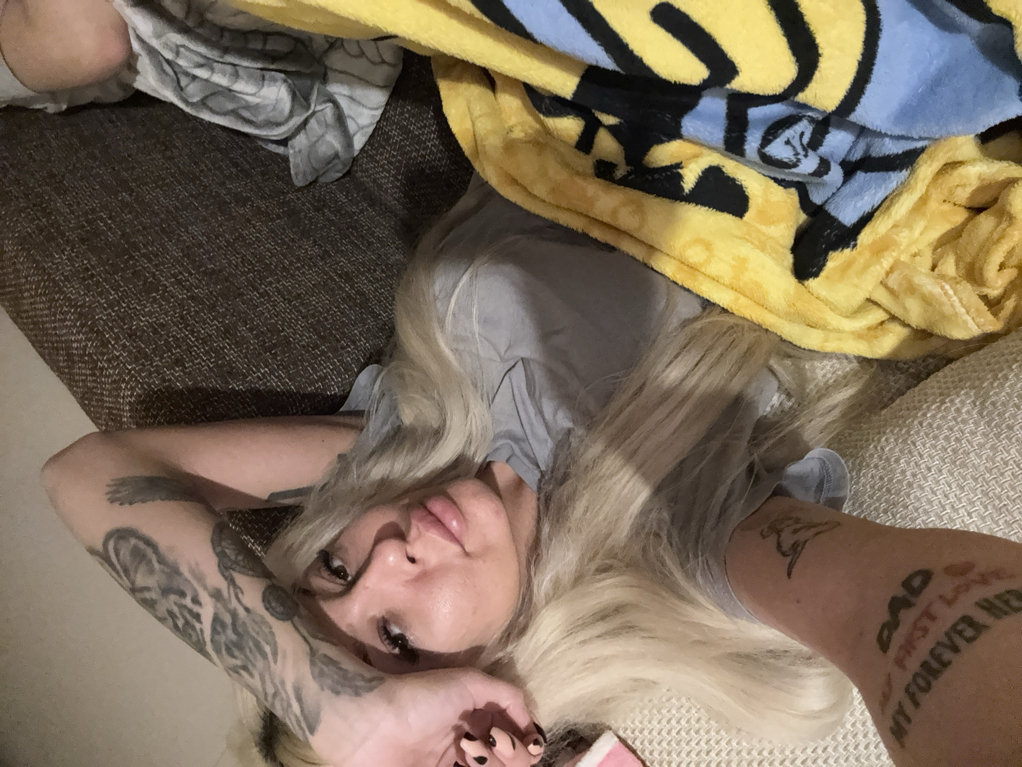 A woman with long blonde hair lies on a brown and beige patterned sofa. A blanket with a yellow and blue pattern is partially covering her. Her arms are bent with her hands near her face. There are tattoos on both forearms. The photo appears to be taken indoors. The location is Unknown, Unknown.