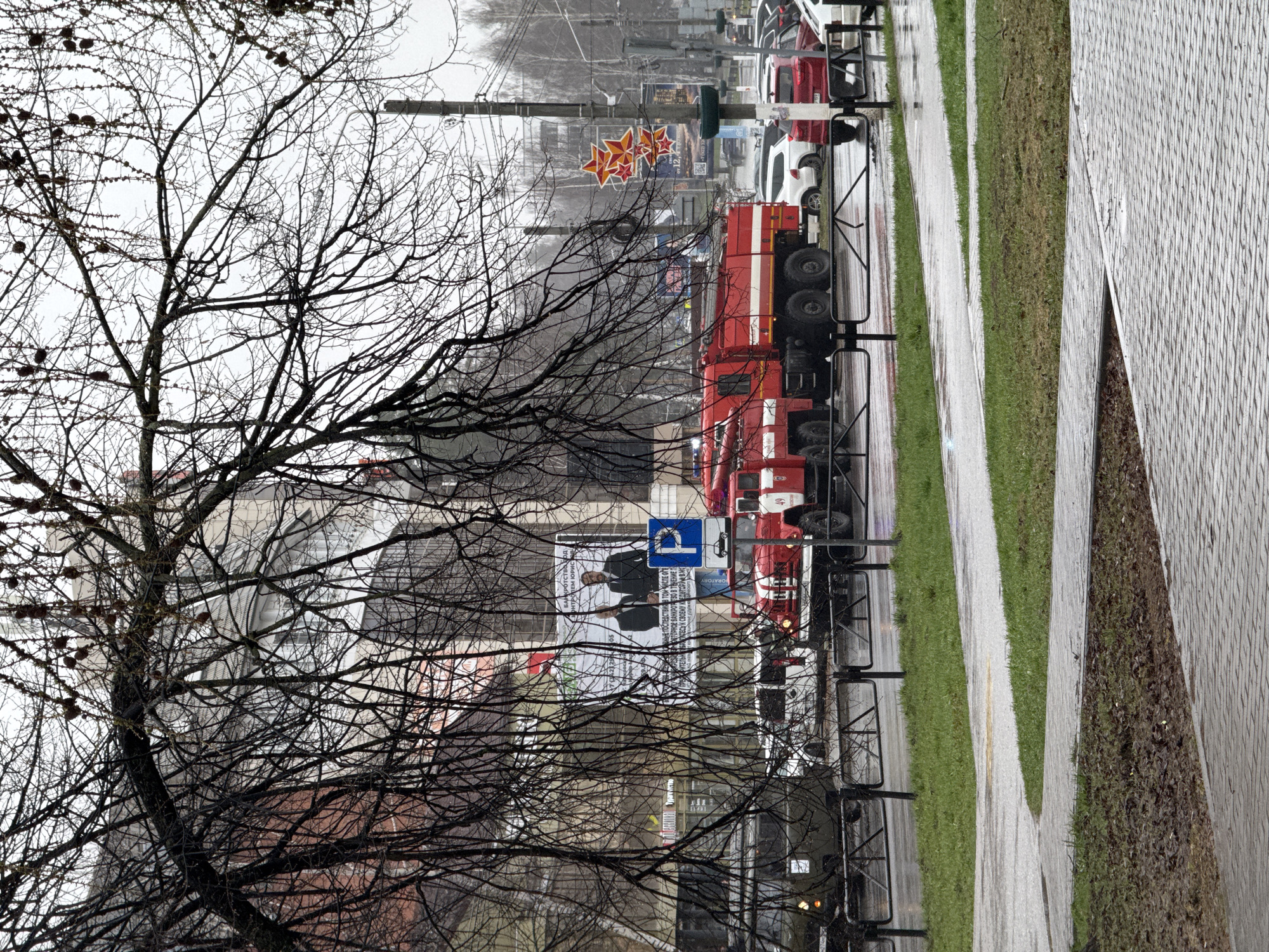 This image captures a street scene in Chaikovsky, Russia, on an overcast and wet day, likely in late autumn, winter, or early spring, given the bare branches of the trees and the moist ground. The perspective looks across a multi-lane road, showing a mix of urban and natural elements.
In the foreground, a prominent, leafless tree with intricate dark branches occupies the left side of the frame. To the right, a wide paved sidewalk or pedestrian path with parallel stripes of grass runs along the side of the street, all appearing damp from recent rain or melting snow.
On the street, a large red fire truck with white stripes is a central subject, positioned amidst other vehicles, including red and white cars. A blue parking sign with a white 'P' and a car icon is clearly visible above the fire truck. Further down the street, suspended wires and utility poles are visible, and a cluster of decorative red stars hangs above the roadway, a common sight in Russian cities, often associated with Victory Day or other patriotic celebrations. A person can be vaguely seen near the fire truck.
In the background, beyond the street, are buildings, one featuring a large billboard or banner.
Visible text from the banner includes: "ПЕМ ЮРИС" (likely a company name, "PEM YURIS"), parts of phrases like "АВНЫЕ ПОСЛЕДСТВИЯ В ТОМ ЧИСЛЕ ОГ" (possibly related to legal consequences), "ОСТОРНОЕ БАНКРОТСТВО В ТЕЧЕНИЕ П" ("careful bankruptcy during..."), and "АТИТЕСЬ К СВОЕМУ КРЕДИТОРИИ В МО" ("contact your creditor in..."). These phrases strongly suggest the banner advertises legal or financial services, possibly related to bankruptcy. Other fragmented text includes "BORATORY" and the number "49". The 'P' on the blue parking sign is also visible.