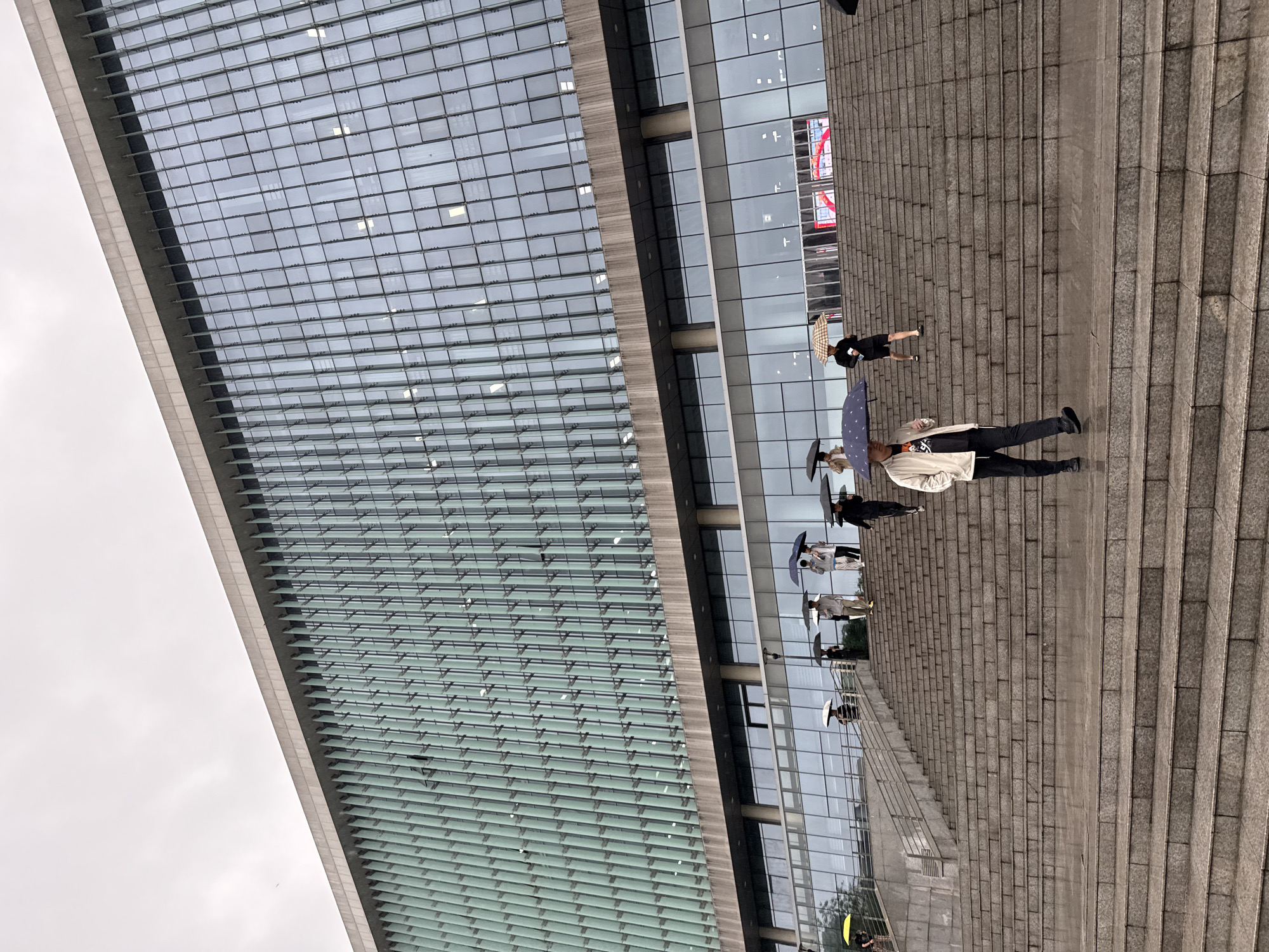 The image displays an architectural outdoor scene featuring a modern building, a grand staircase, and several people under an overcast sky in Jiangjin, China.
On the left side of the frame, a tall, contemporary building rises upwards. Its facade is composed predominantly of glass panels arranged in a grid pattern, with horizontal louvers or shading elements integrated across sections of the windows. The glass reflects the grey sky and indistinct shapes.
To the right, a wide, light grey stone staircase ascends diagonally across the image. Several individuals are present on these steps, generally walking uphill. A notable detail is that many of the people are holding open umbrellas, suggesting either rain or protection from the sun. One person near the bottom right, dressed in dark attire, holds a light-colored, patterned umbrella. Further up, another individual in a light jacket and dark pants carries an open, dark blue umbrella. Other people with umbrellas are visible higher on the stairs. One person near the building's entrance appears to be holding up a phone or camera, possibly taking a photo or recording.
The sky, visible in the upper left, is uniformly light grey and overcast, consistent with the use of umbrellas for weather protection. Inside the building, visible through the glass facade, a large screen displays an indiscernible red graphic or text. The scene depicts a public space or the approach to a large modern structure, bustling with activity on a cloudy day.