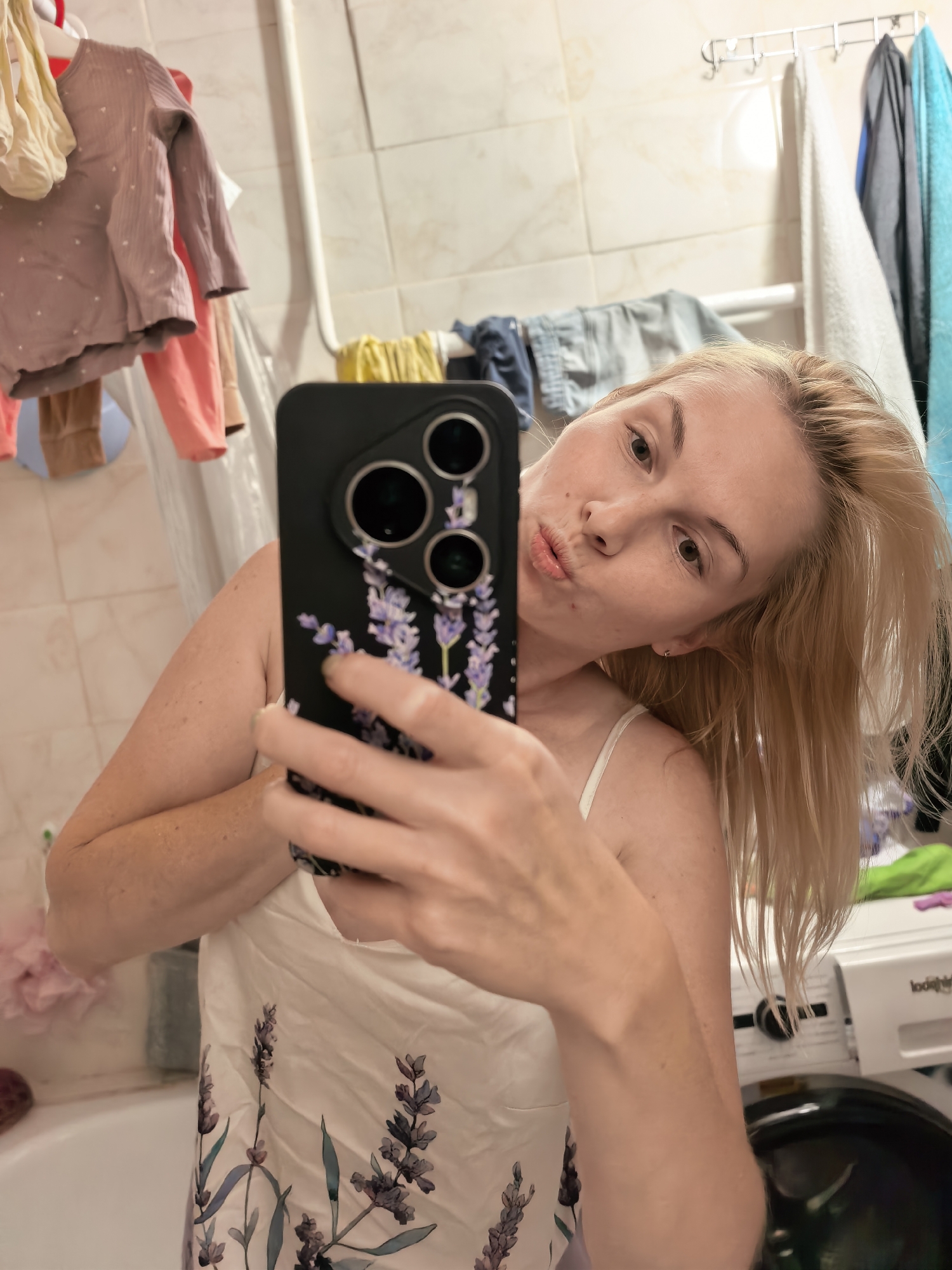 In a bathroom, a blonde woman with fair skin and freckles is taking a mirror selfie. She is wearing a white camisole with a lavender floral pattern and her hair is blonde and medium-length. She is holding up a smartphone with a black case adorned with a lavender design and three camera lenses. The background shows a tiled wall, a clothes rack with laundry hanging on it, and a washing machine. The scene appears to be in a residential bathroom.
