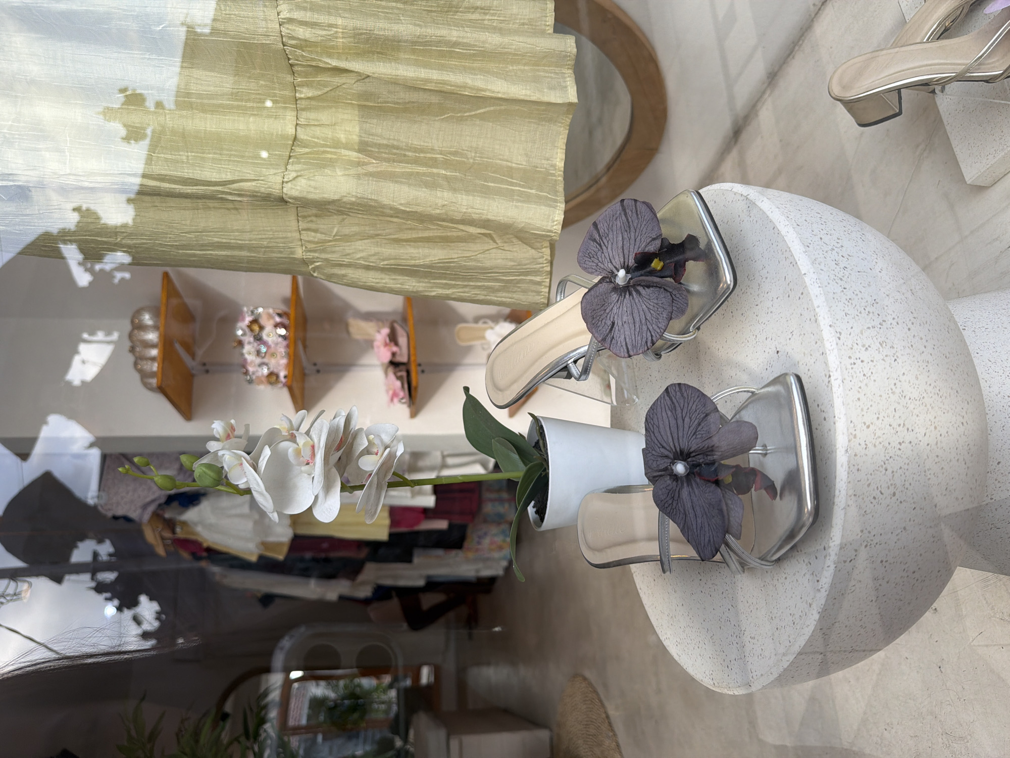 The image shows a display in what appears to be a fashion boutique. Two pairs of women's sandals with silver straps and a single dark gray flower on each are displayed on small white terrazzo-style pedestals. A potted plant with white orchids is positioned between the sandals. The background reveals a clothing rack with garments and a round mirror. The reflective surfaces suggest the display is behind glass, possibly in a storefront. Given the setting, it is presumed the location is in Kuta, Indonesia, as specified.